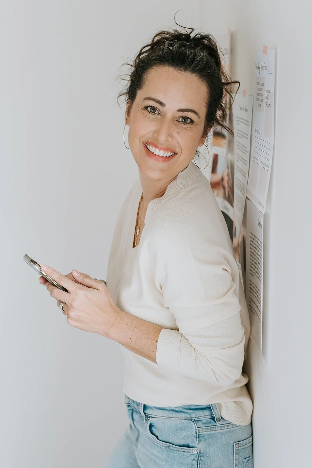 Smiling woman with dark curly hair using a smartphone, standing against a white wall decorated with papers.