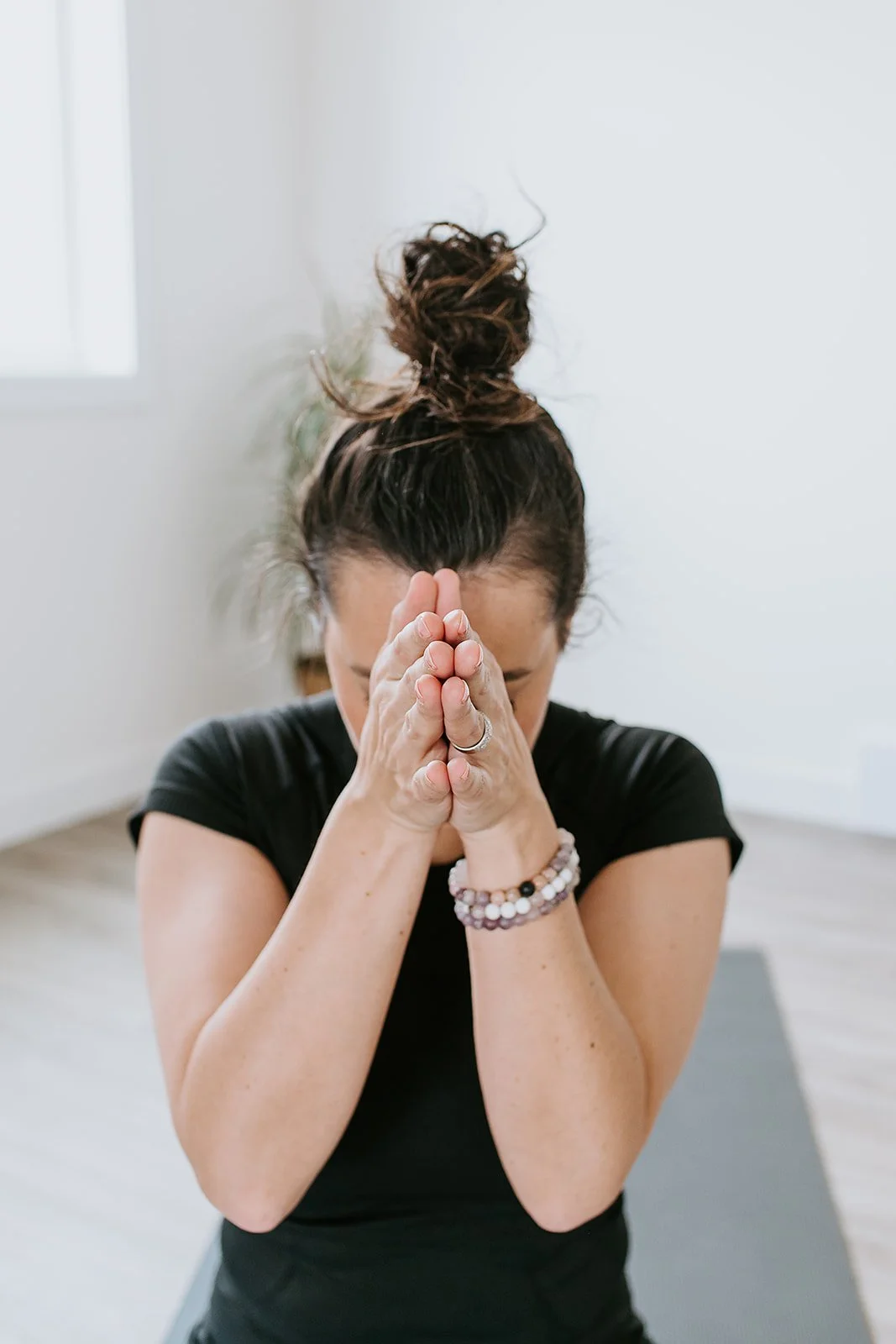 Woman practicing yoga or meditation, bowing with hands pressed together in prayer pose, wearing black shirt and beaded bracelets, in a bright room with wooden flooring