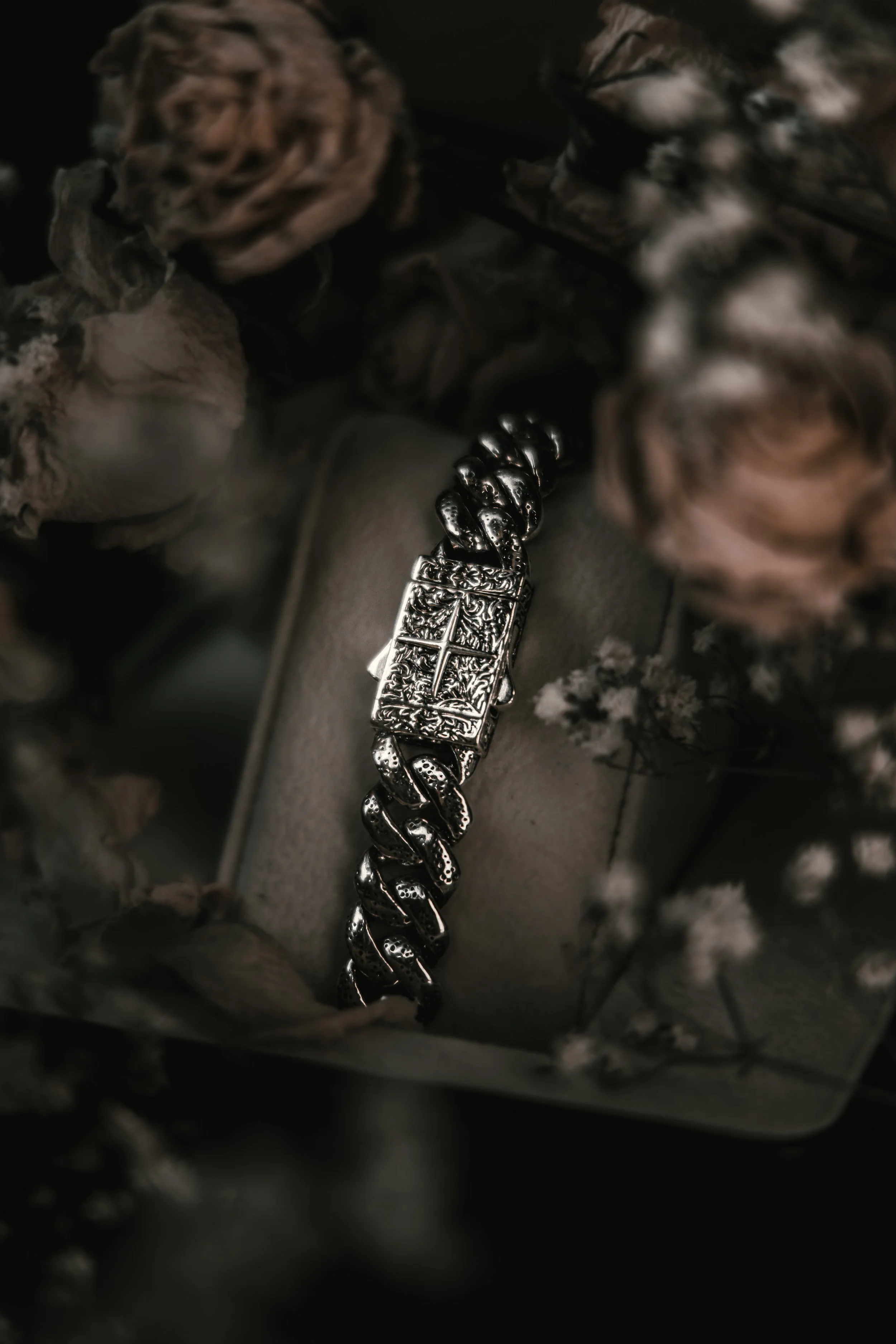 A silver bracelet with a detailed cross charm surrounded by dried flowers on a neutral surface.