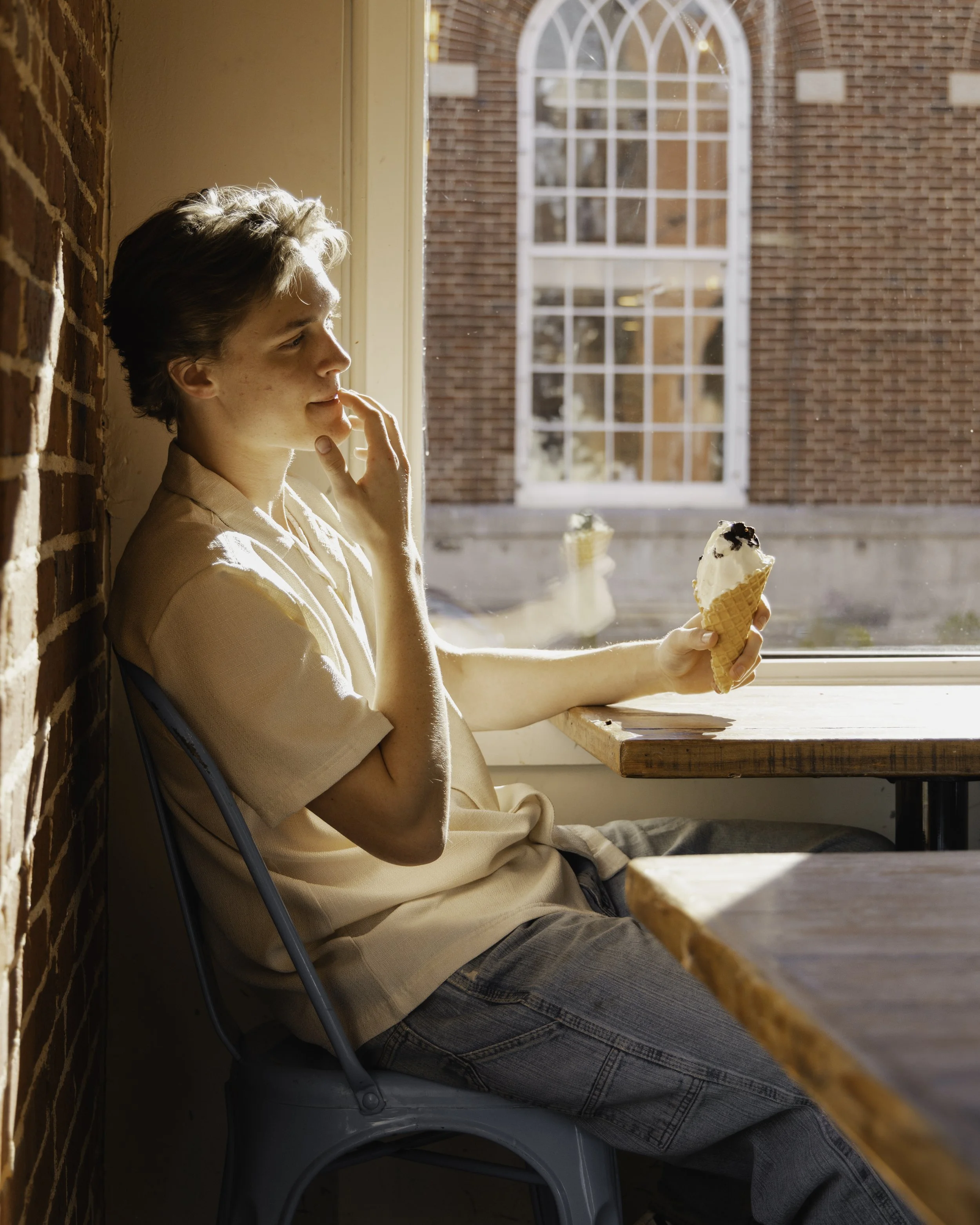A young man with short, wavy hair, wearing a beige polo shirt and jeans, sitting by a large window in a cafe, holding an ice cream cone, with sunlight streaming in.