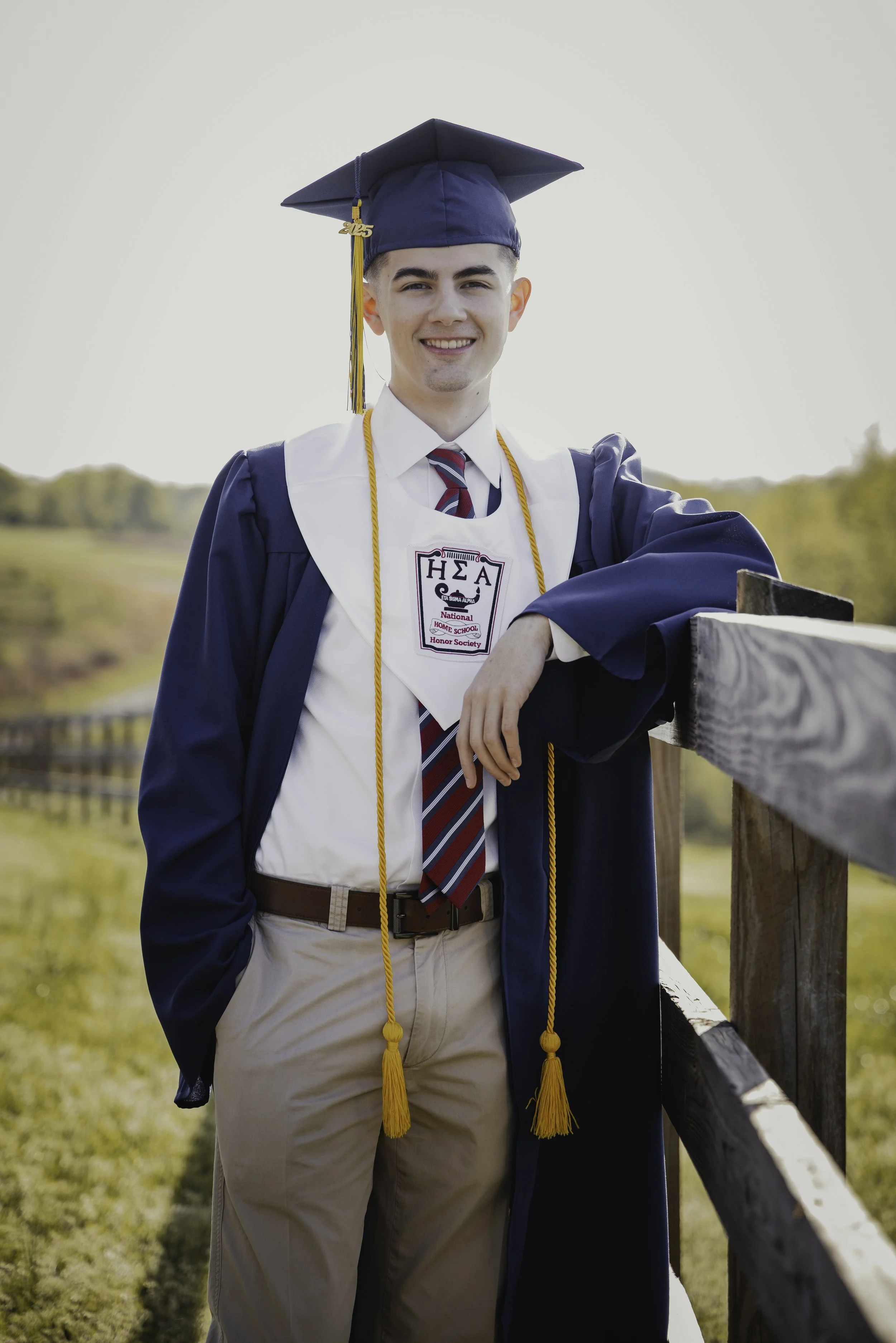 Young man in graduation cap and gown smiling outdoors, leaning on a wooden railing with a scenic background.