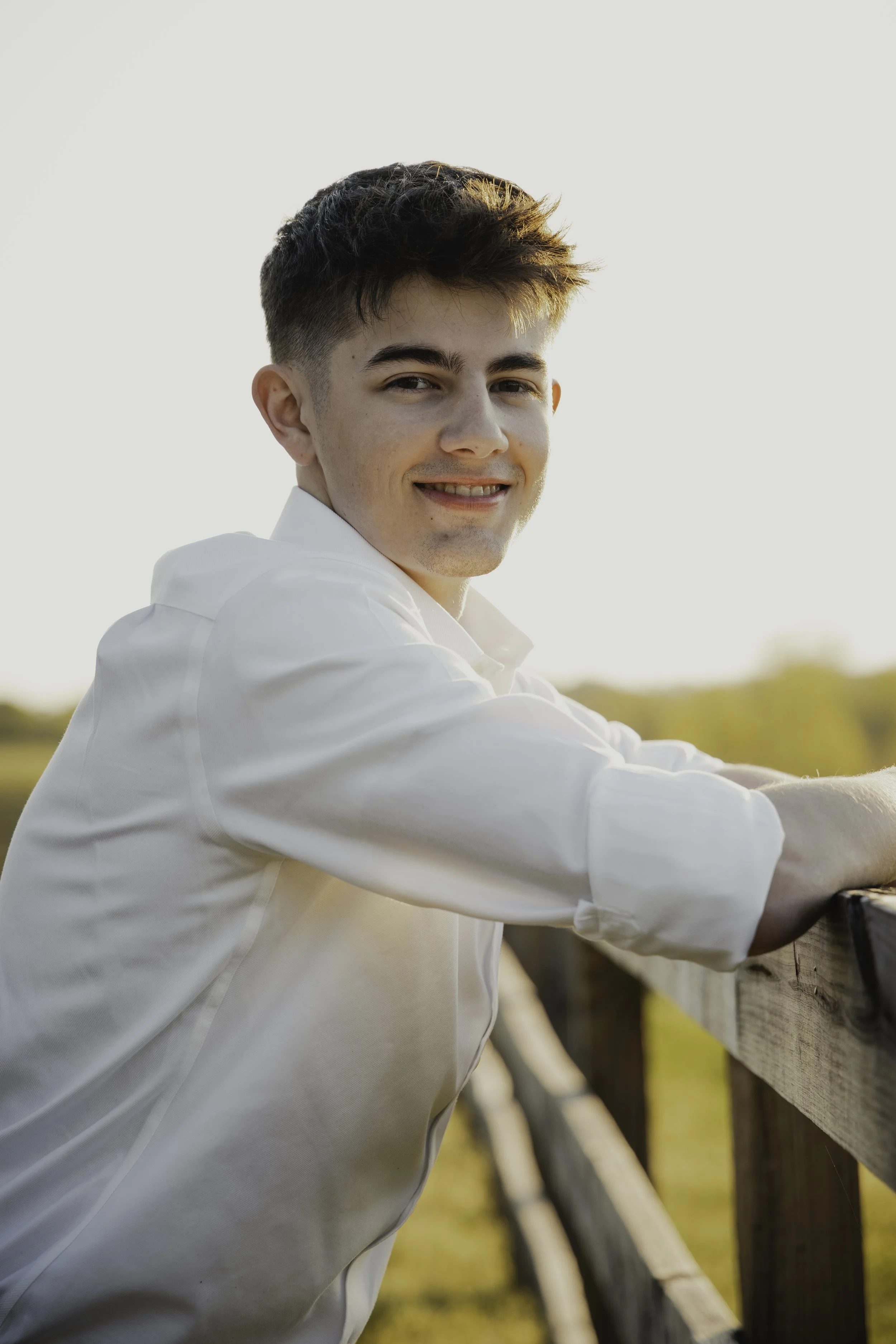 A young man in a white shirt leaning on a wooden fence outdoors at sunset, smiling at the camera.
