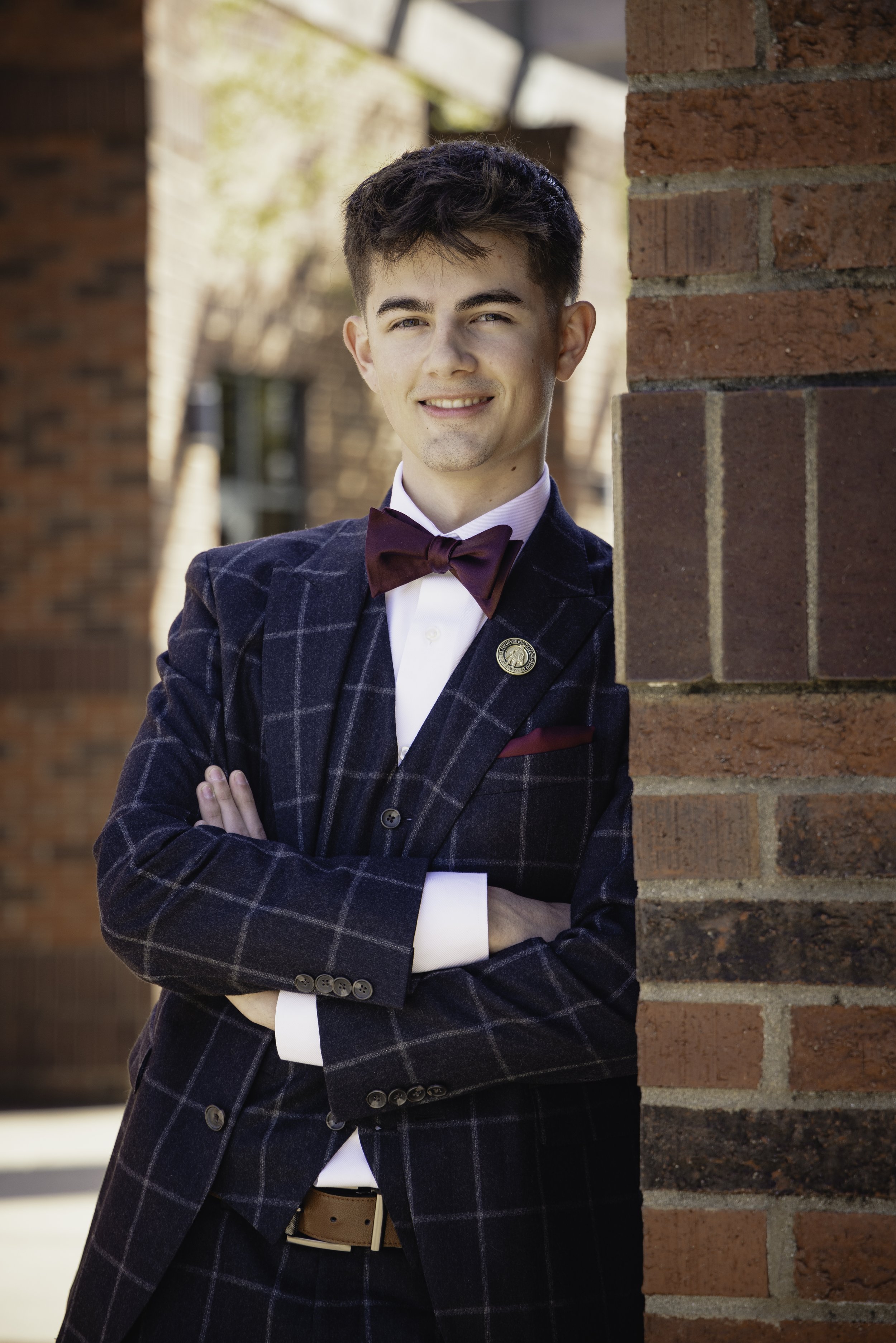 A young man wearing a dark plaid suit, white shirt, and maroon bow tie, standing outdoors next to a brick wall, smiling with arms crossed.