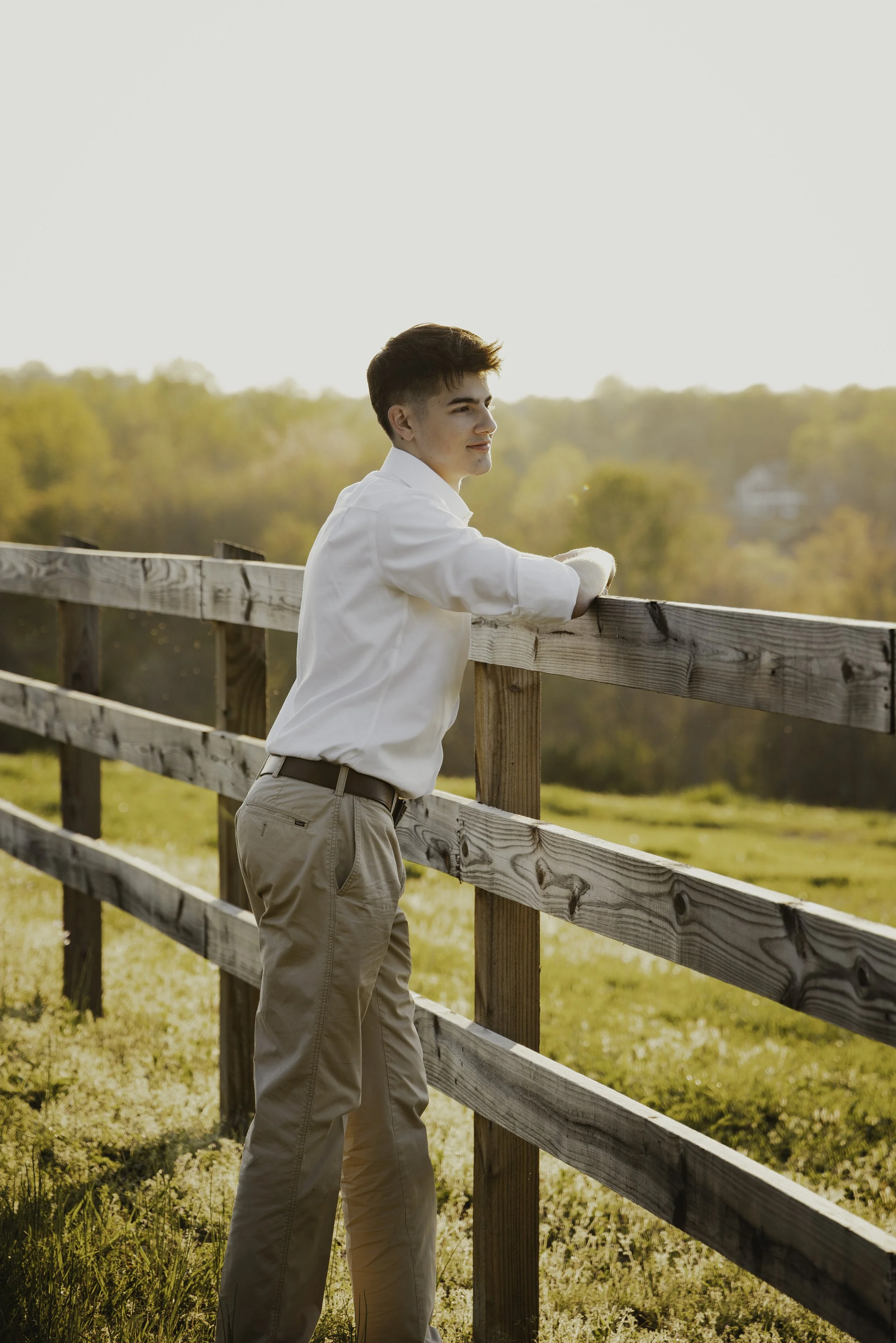 A young man in a white shirt and beige pants stands near a wooden fence, gazing at a sunset over a landscape with trees and open land.