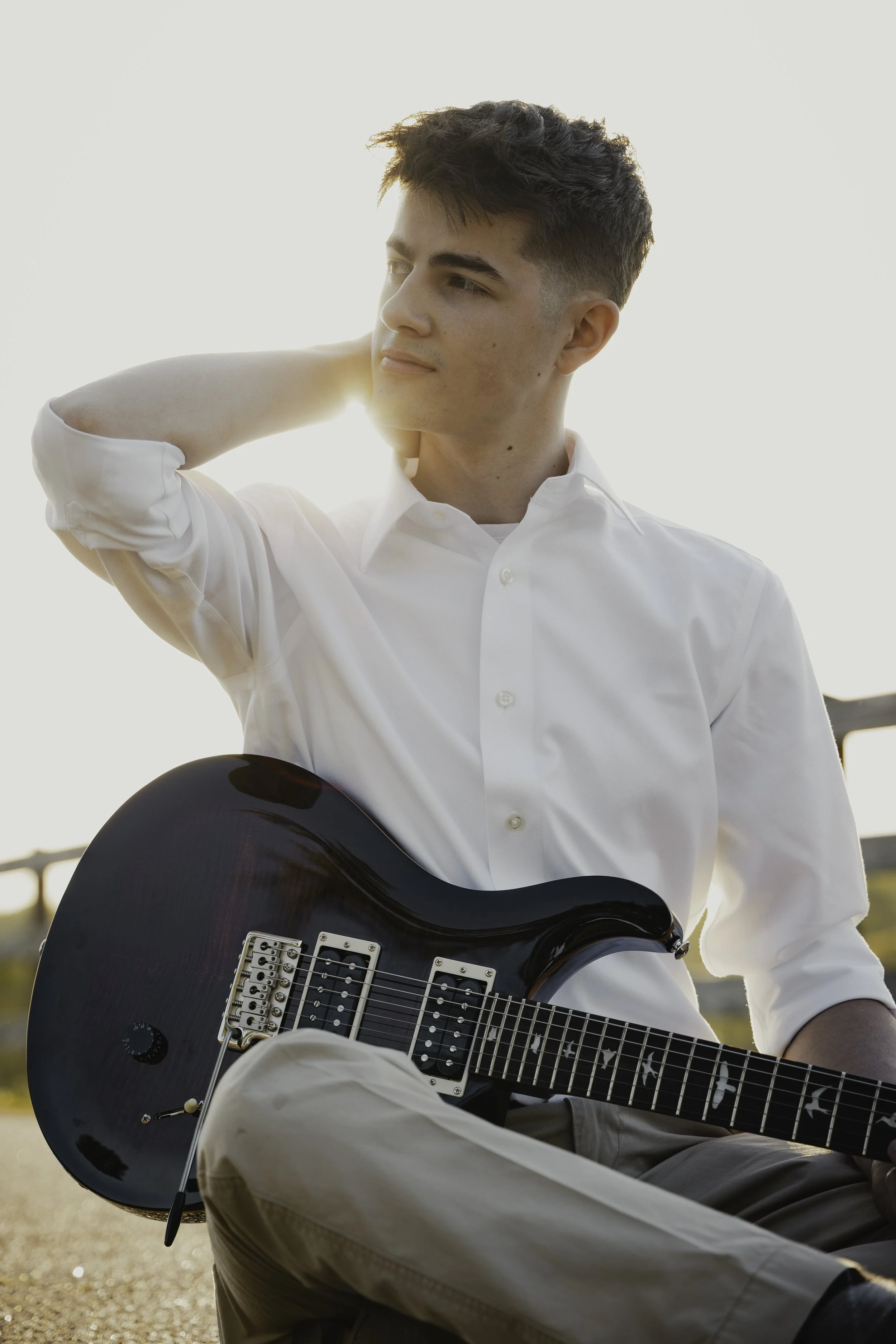 A young man with dark hair wearing a white shirt, sitting outdoors with a black electric guitar, looking pensively to the side during sunset.