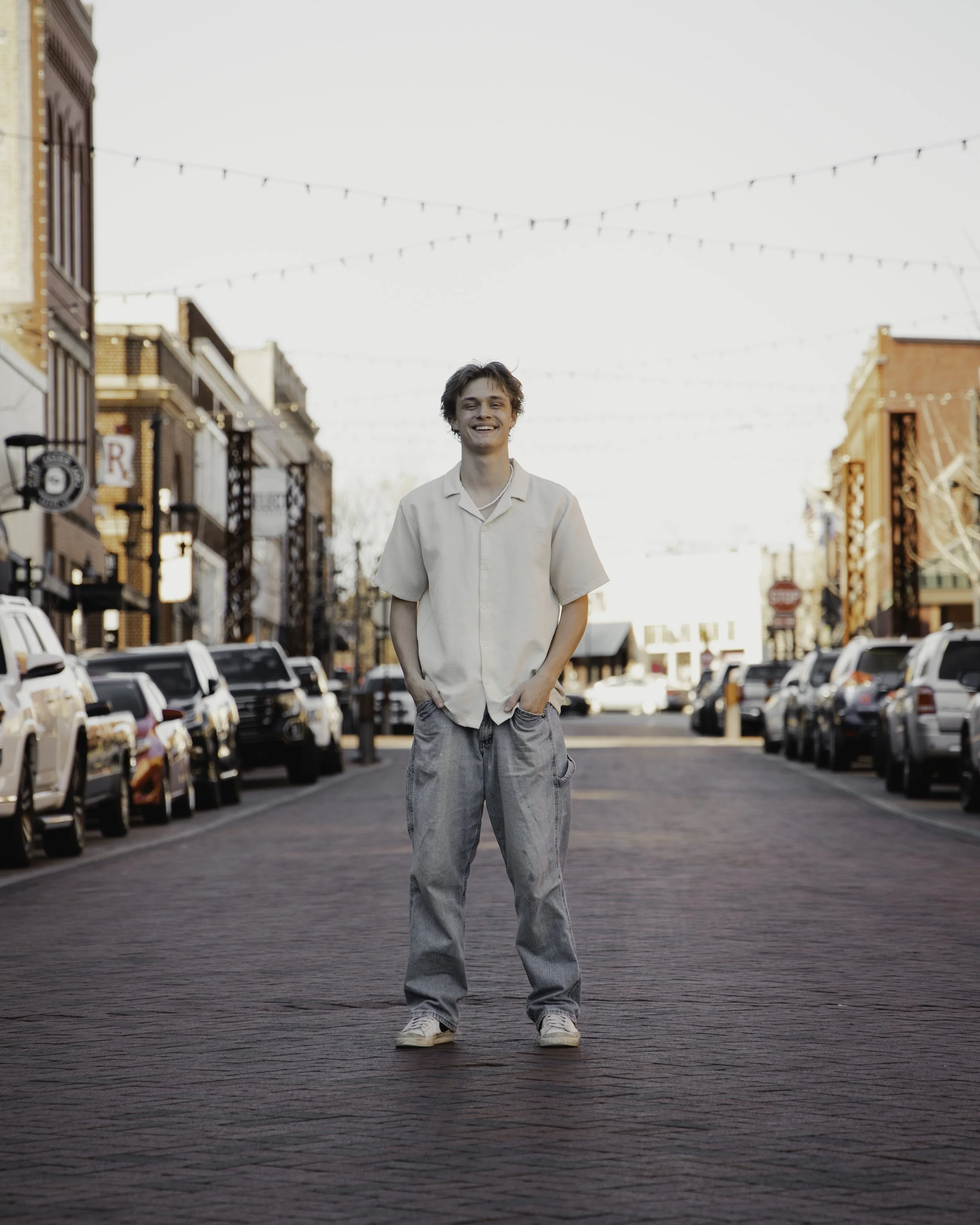 Young man smiling and standing with hands in pockets on a brick street in downtown area with parked cars and buildings, string lights overhead, during daytime.