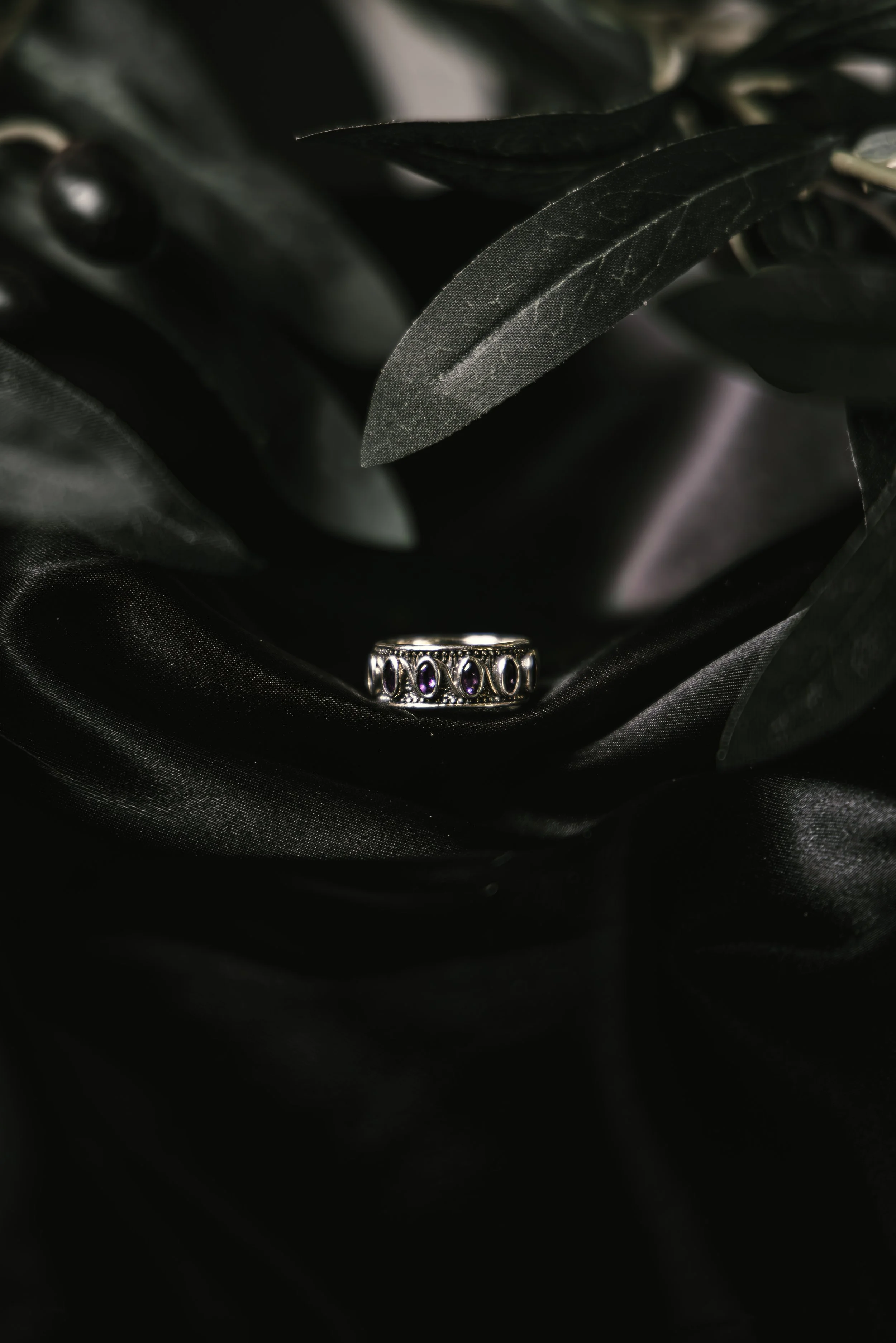 A silver ring with purple gemstones resting on black satin fabric, surrounded by black satin leaves.
