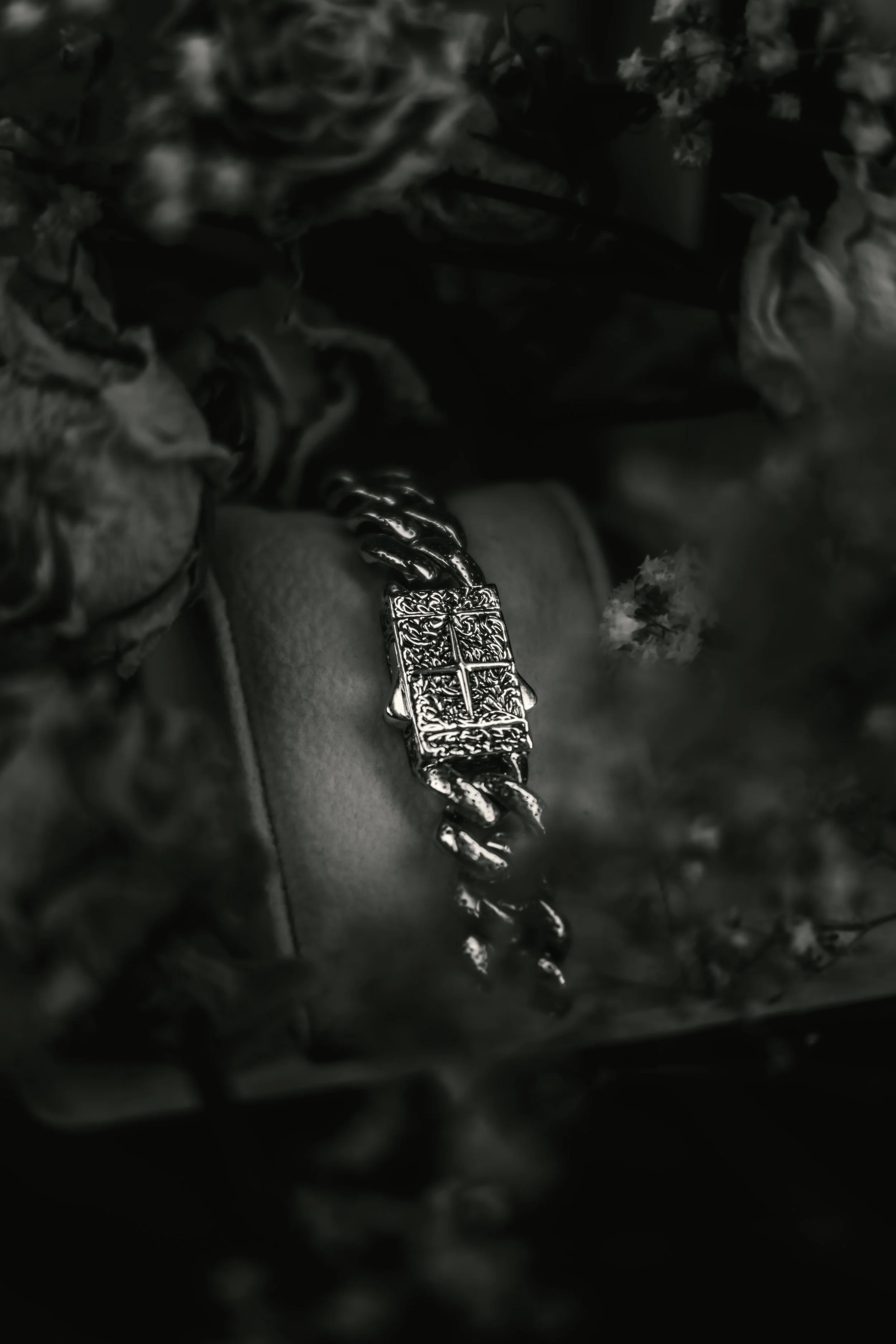 A silver chain bracelet with a rectangular ornate clasp, surrounded by blurred flowers.