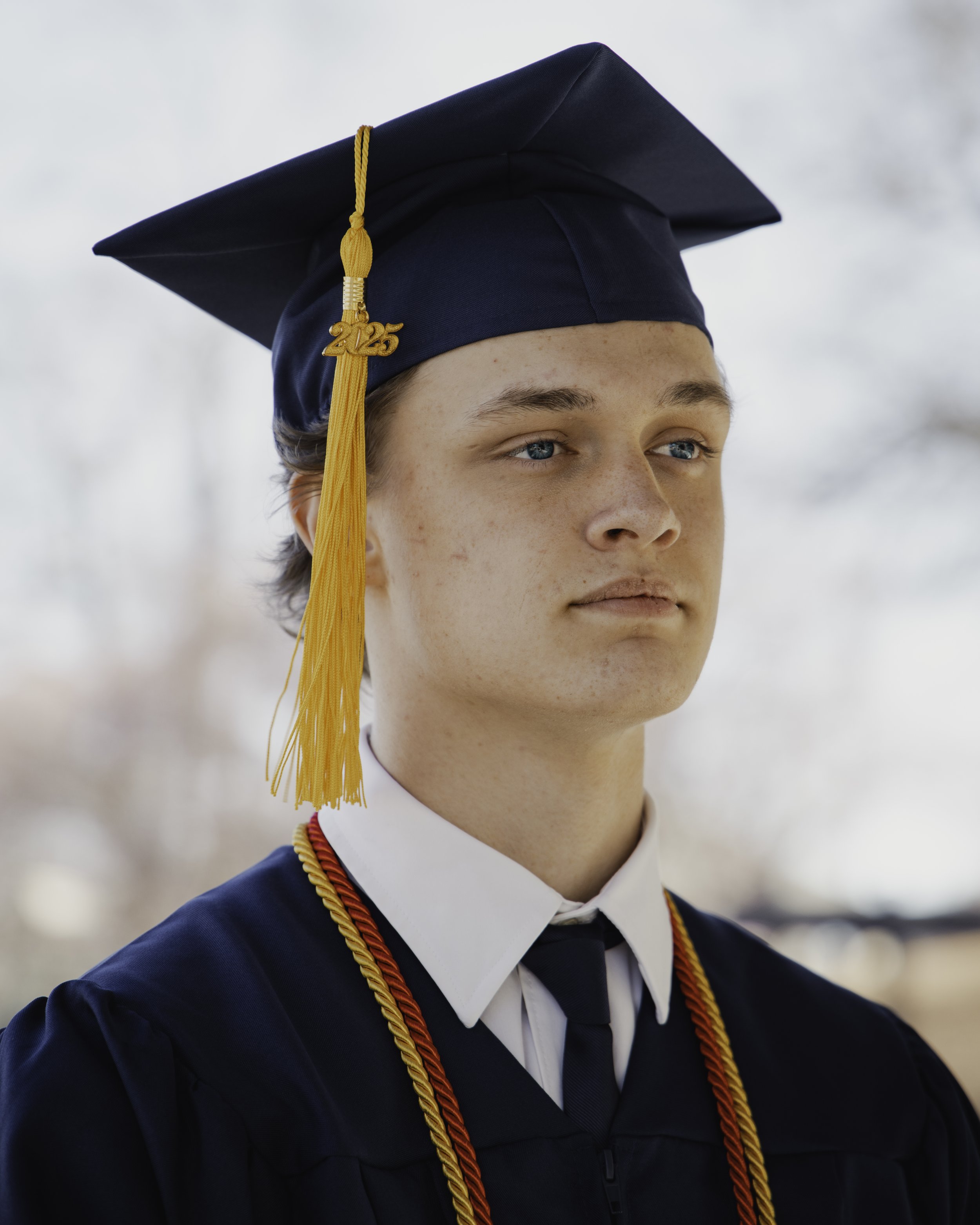 A young man in a graduation cap and gown, wearing a white shirt, black tie, and honor cords, standing outdoors with a blurred background.