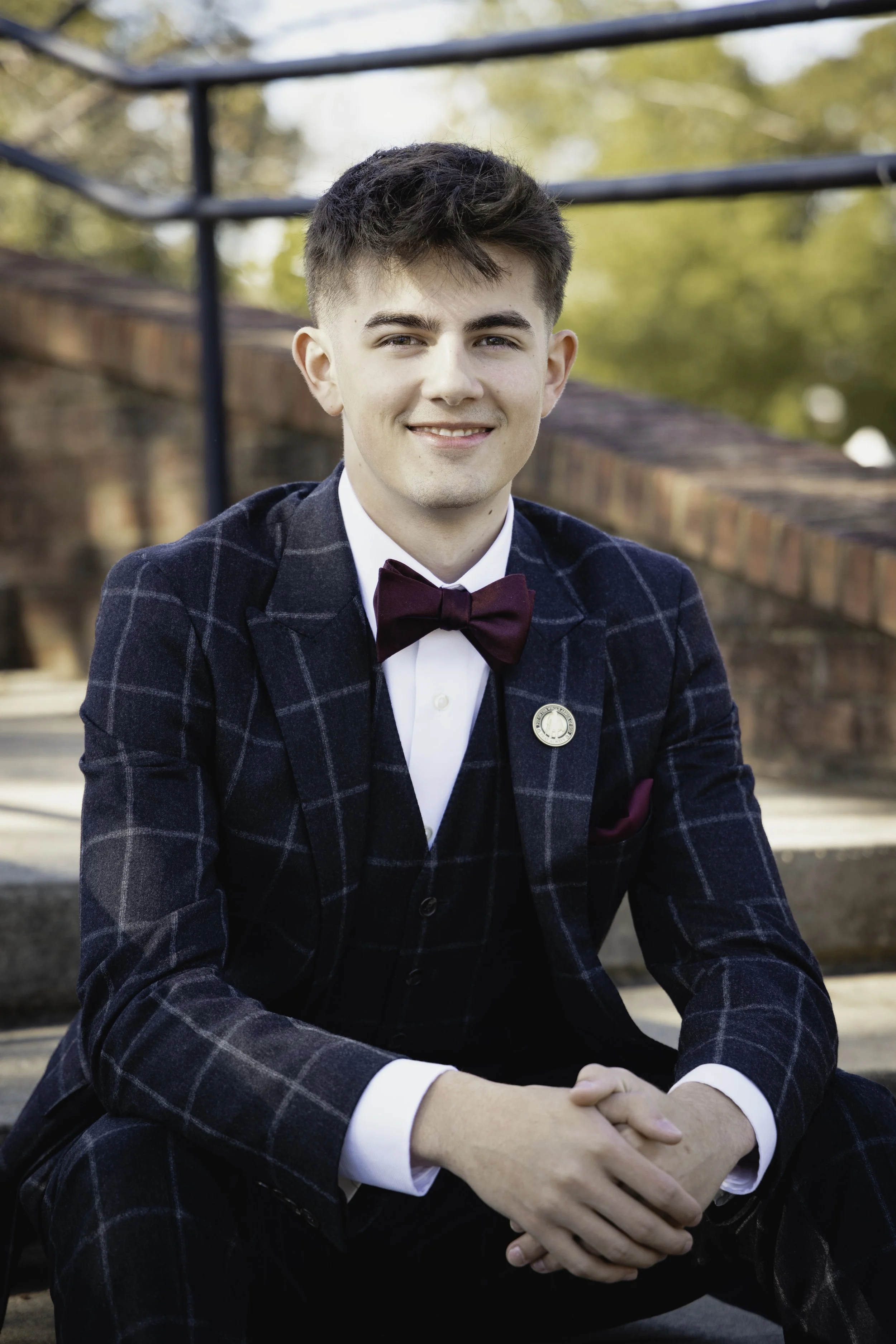 A young man wearing a dark plaid tuxedo with a burgundy bow tie, sitting outdoors on steps with a brick wall and trees in the background.