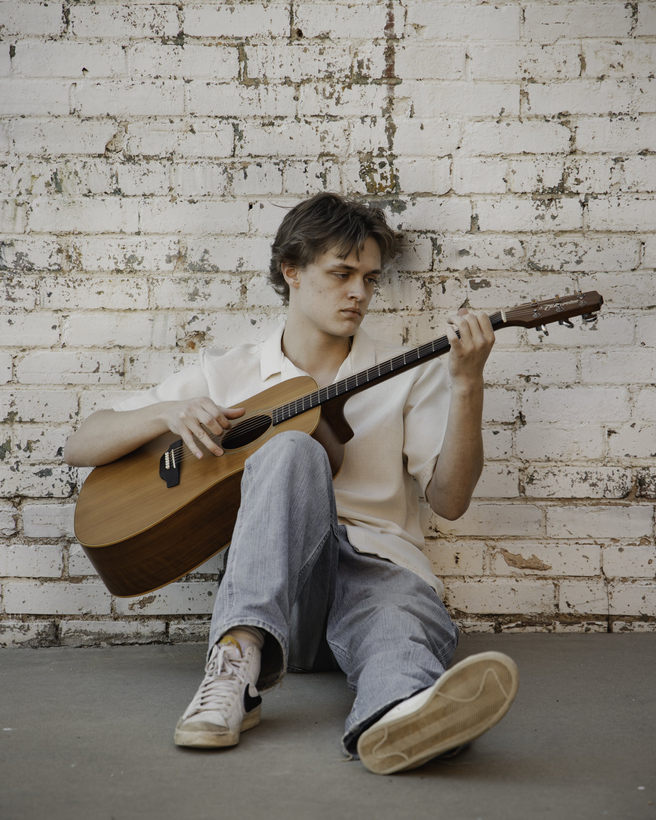 Young man sitting on the ground, leaning against a white brick wall, playing an acoustic guitar.