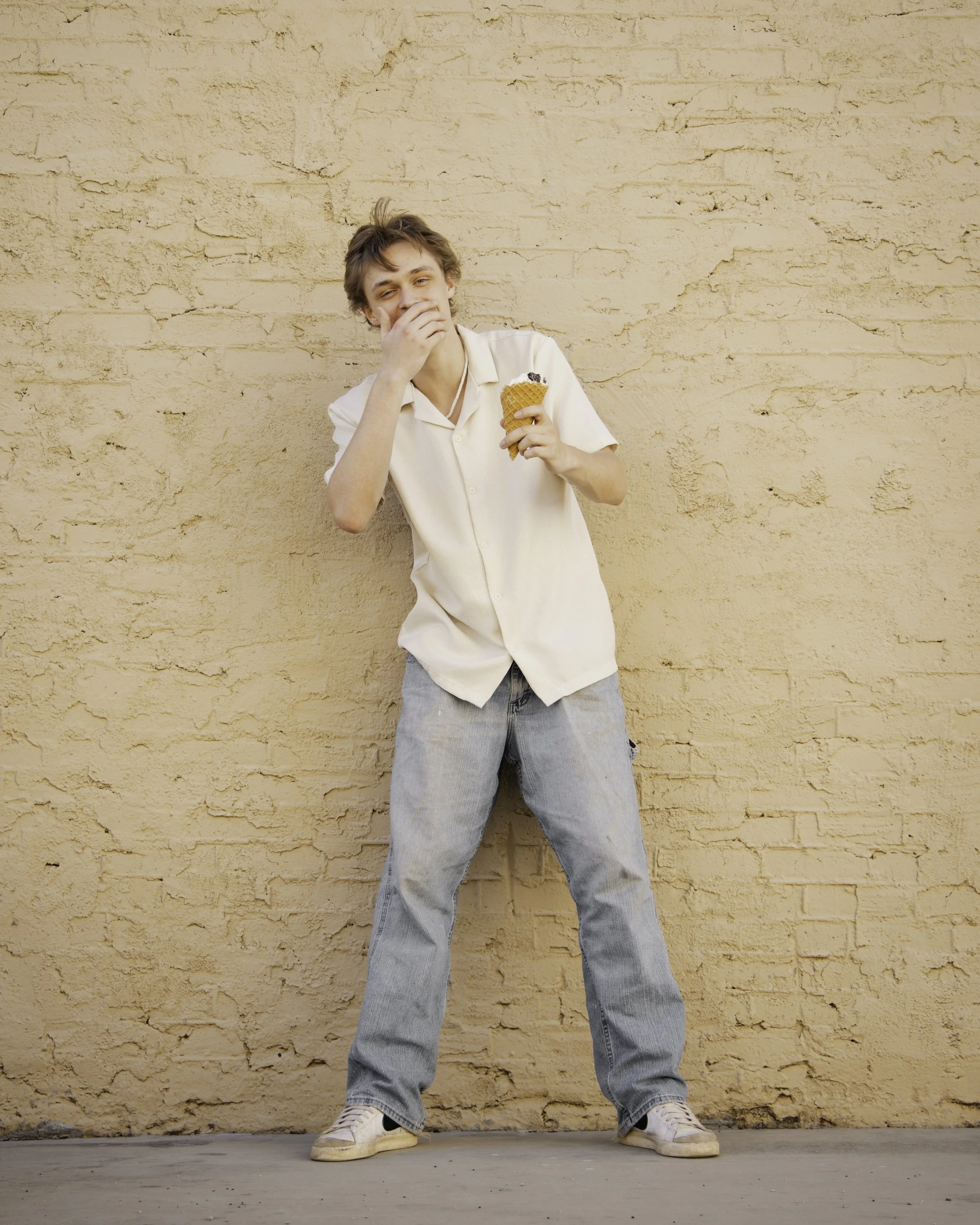 A young man standing against a beige textured wall, laughing while holding an ice cream cone.