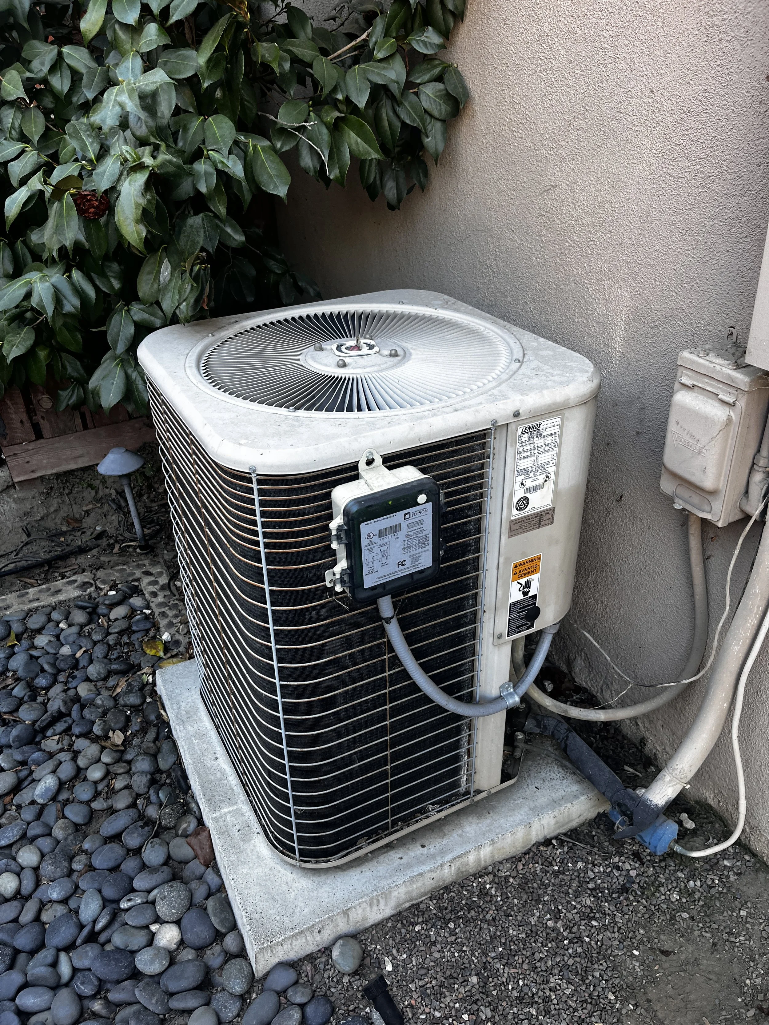 An outdoor air conditioning unit sitting on a concrete pad next to a beige wall and green foliage, with electrical components and piping connected.