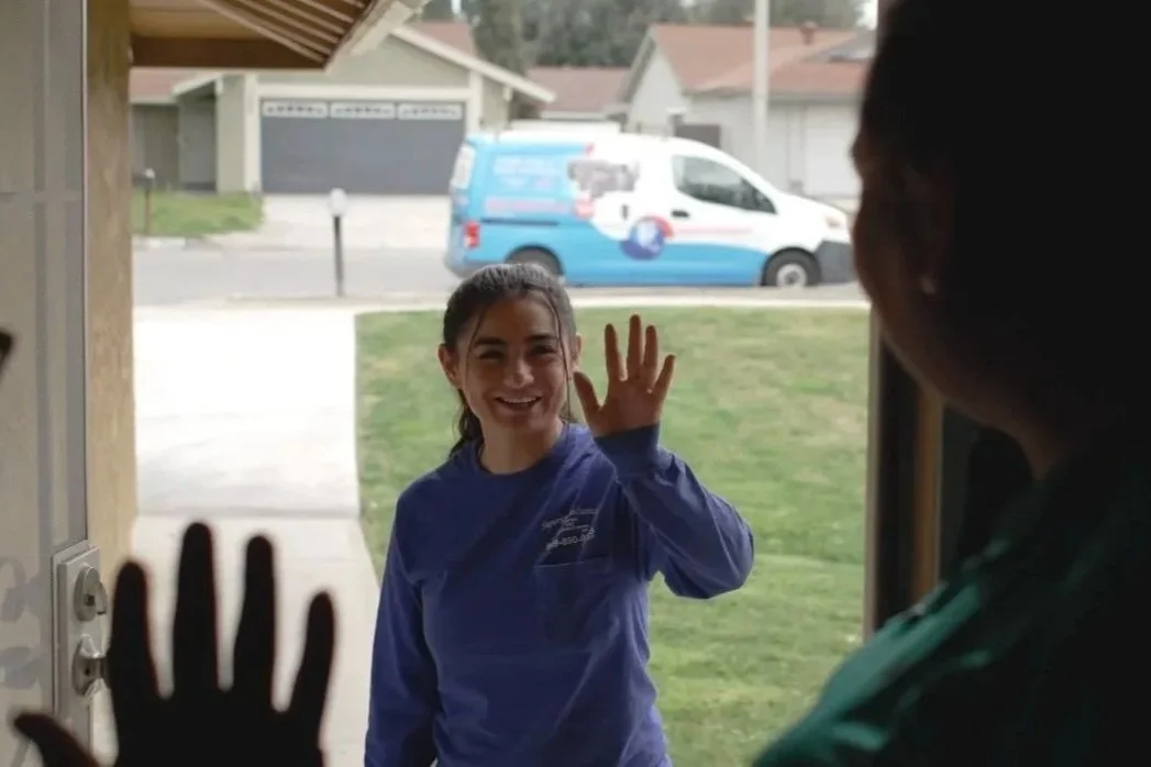 A girl in a blue jacket smiling and waving at a woman through a glass door, outside on a grassy yard with a street and houses in the background.