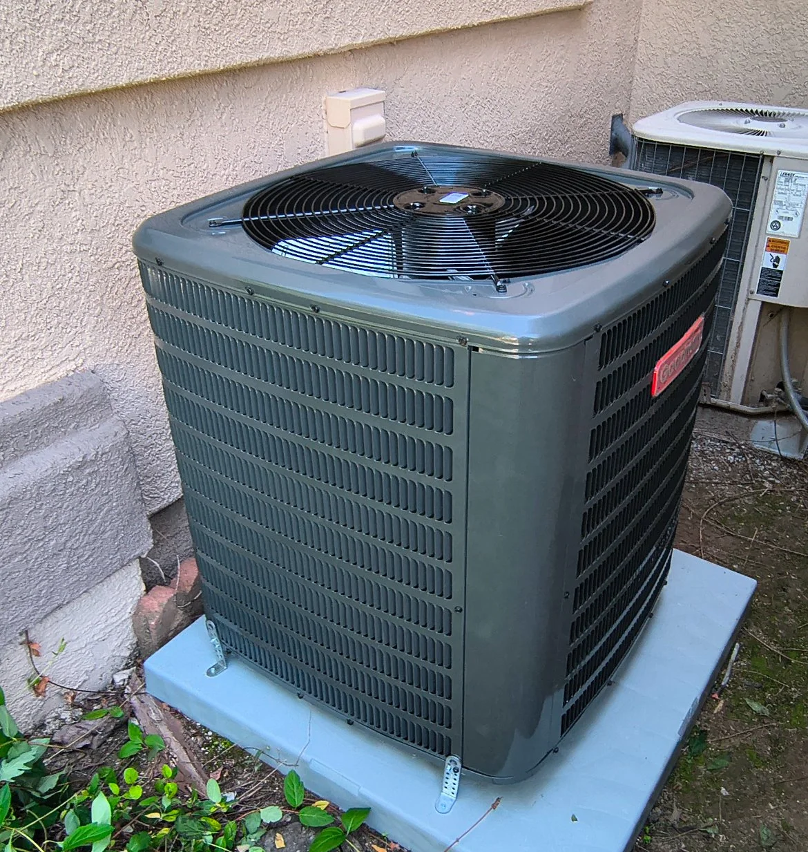 An outdoor air conditioning unit with a large fan on top, situated on a concrete pad beside a textured beige wall.