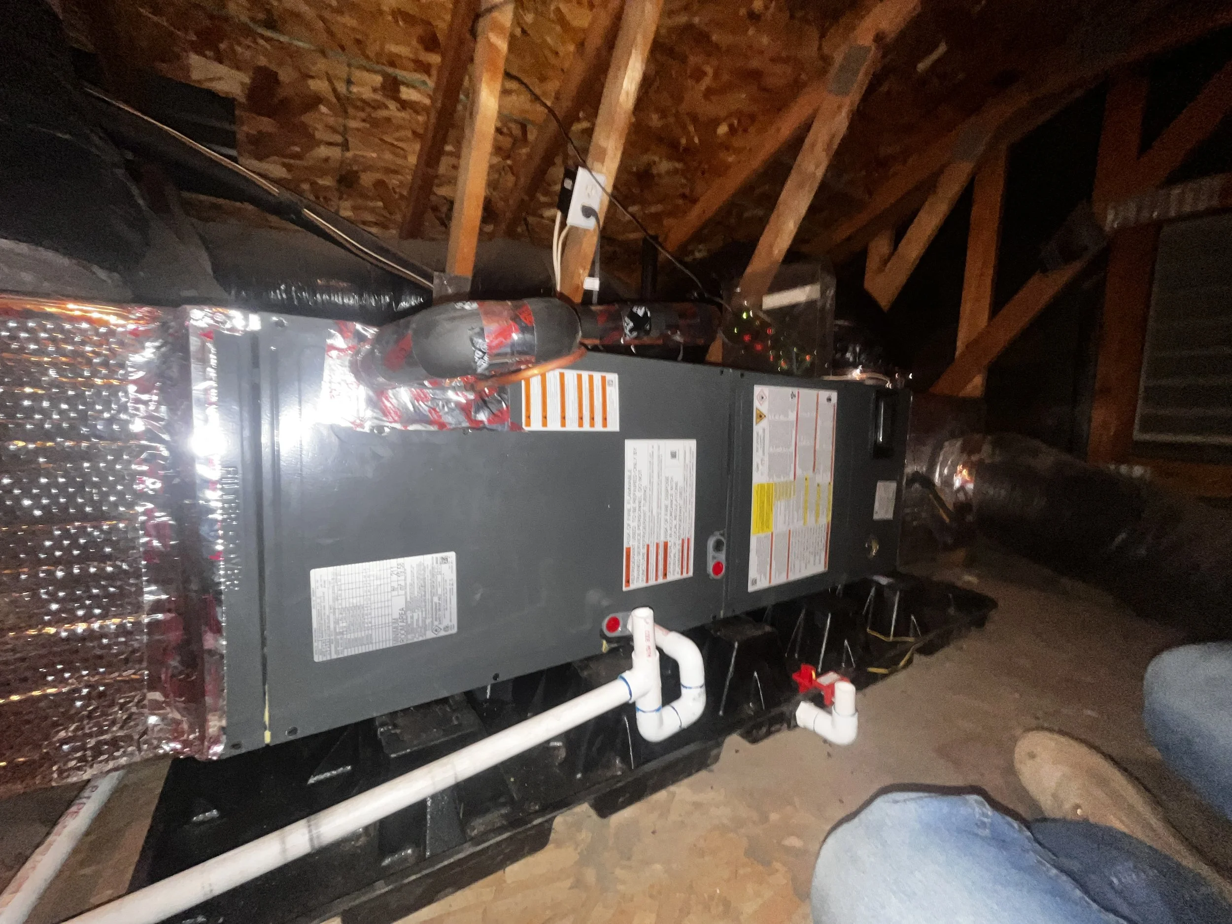 A furnace installed in an attic space with exposed wooden beams, connected to plastic and metal piping, and electrical wiring.