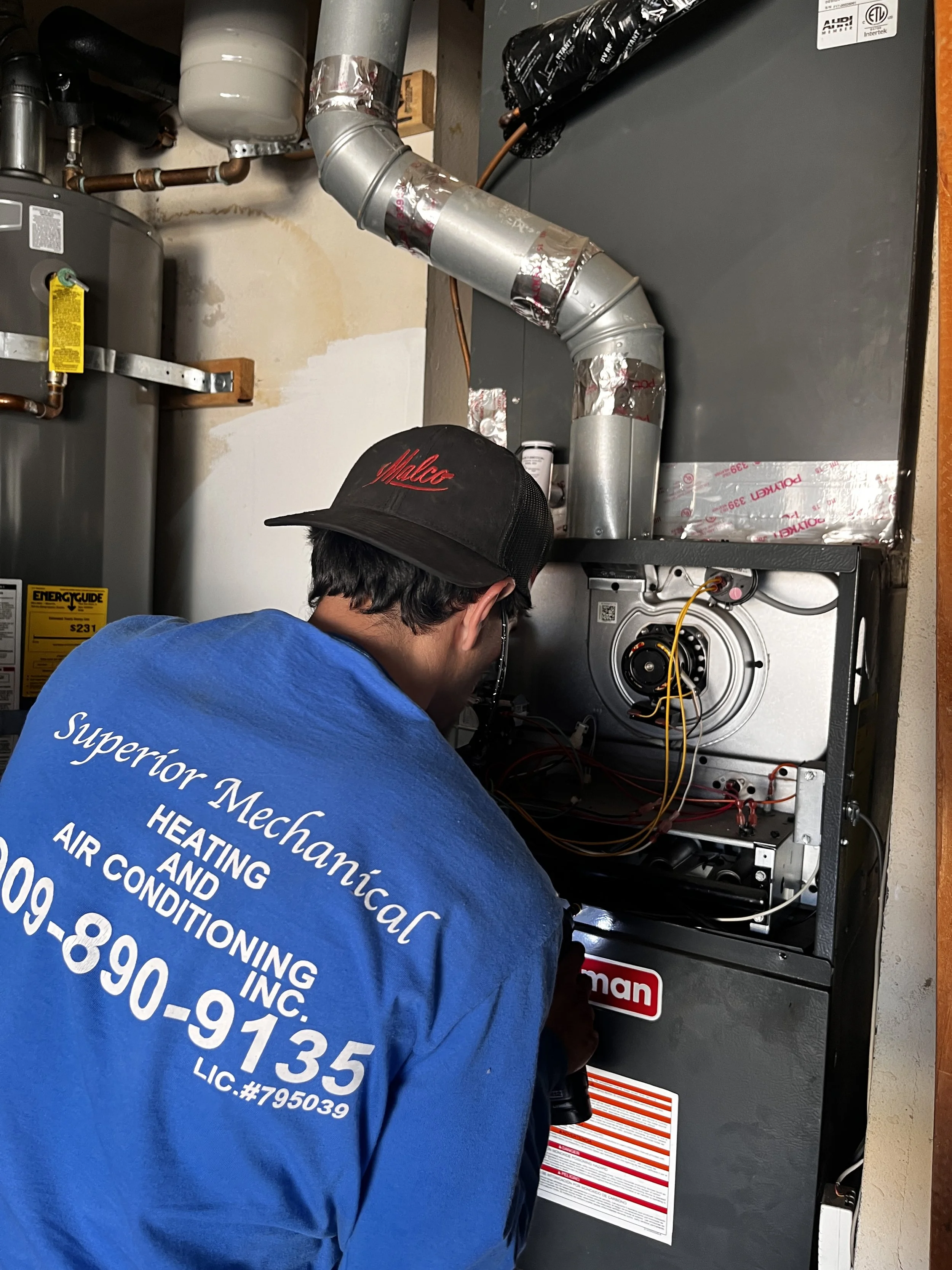 A person working on a furnace or HVAC system, with exposed internal components and ductwork, wearing a blue shirt with HVAC company information, including a phone number and license number, and wearing a black cap.