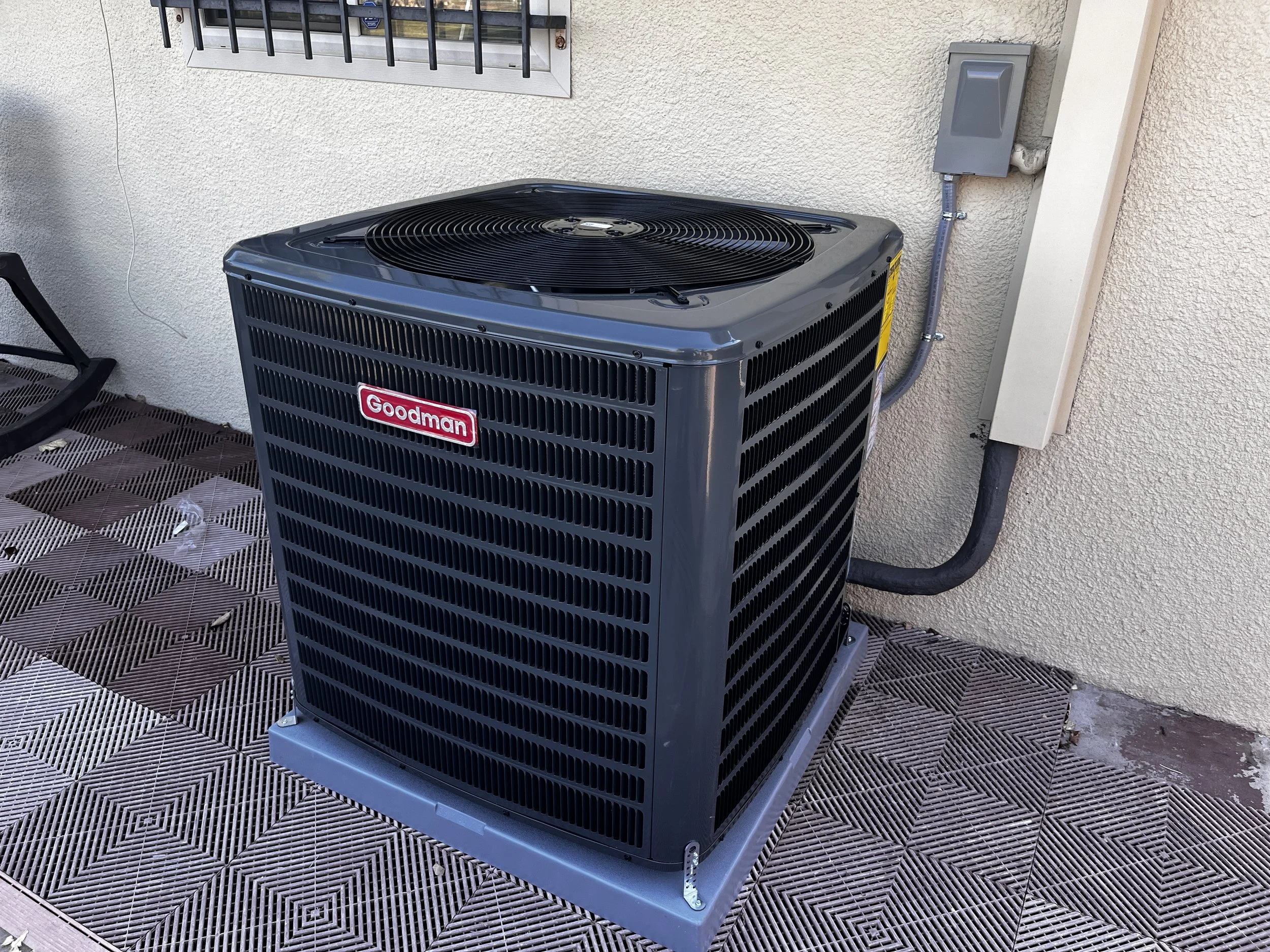 An outdoor view of a Goodman air conditioning unit installed on a patterned metal deck against a textured beige wall. There is an electrical box and a window with security bars above the unit.