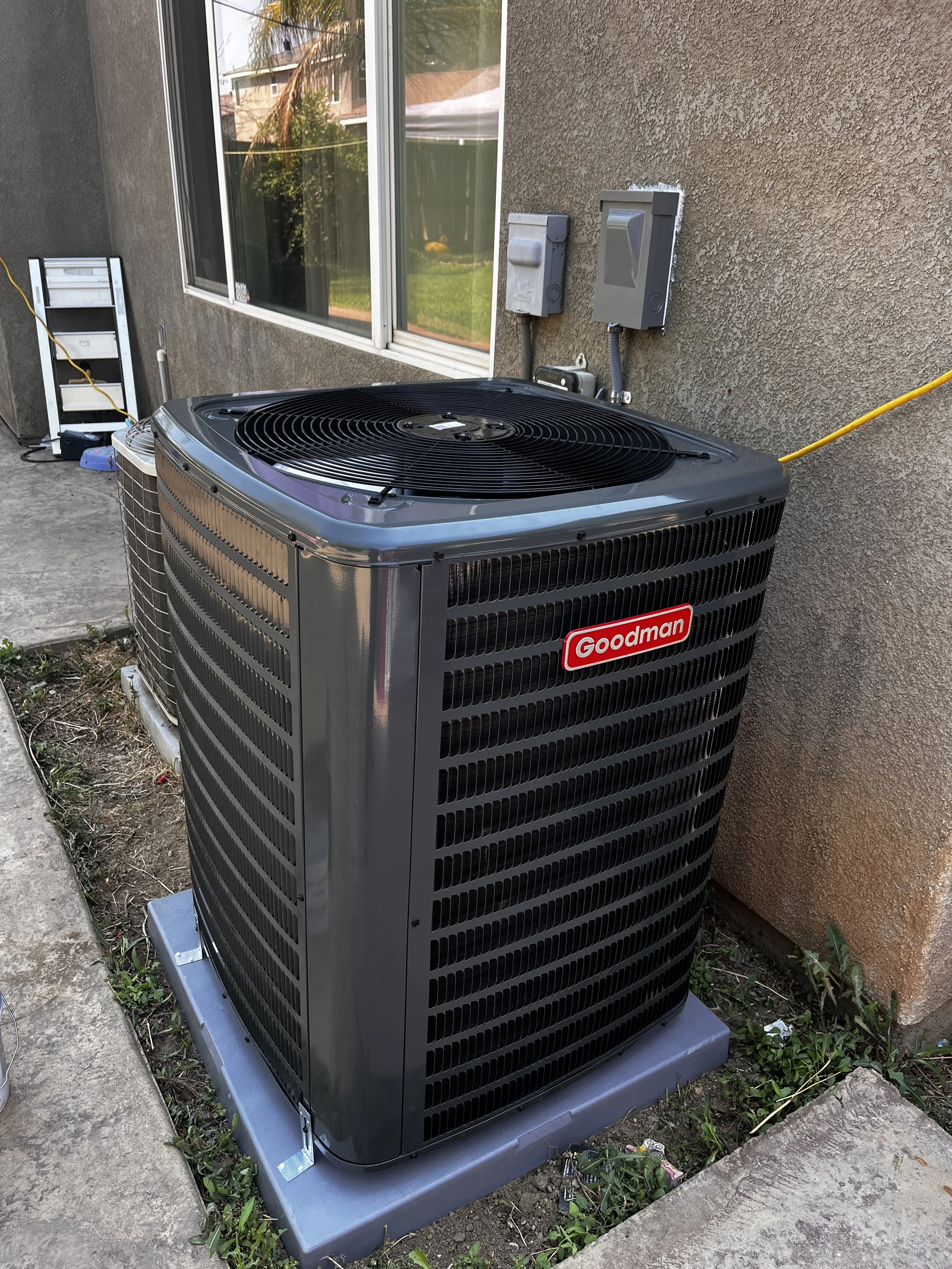 An outdoor Goodman air conditioning unit installed next to a building wall, with electrical connections and a window visible in the background.