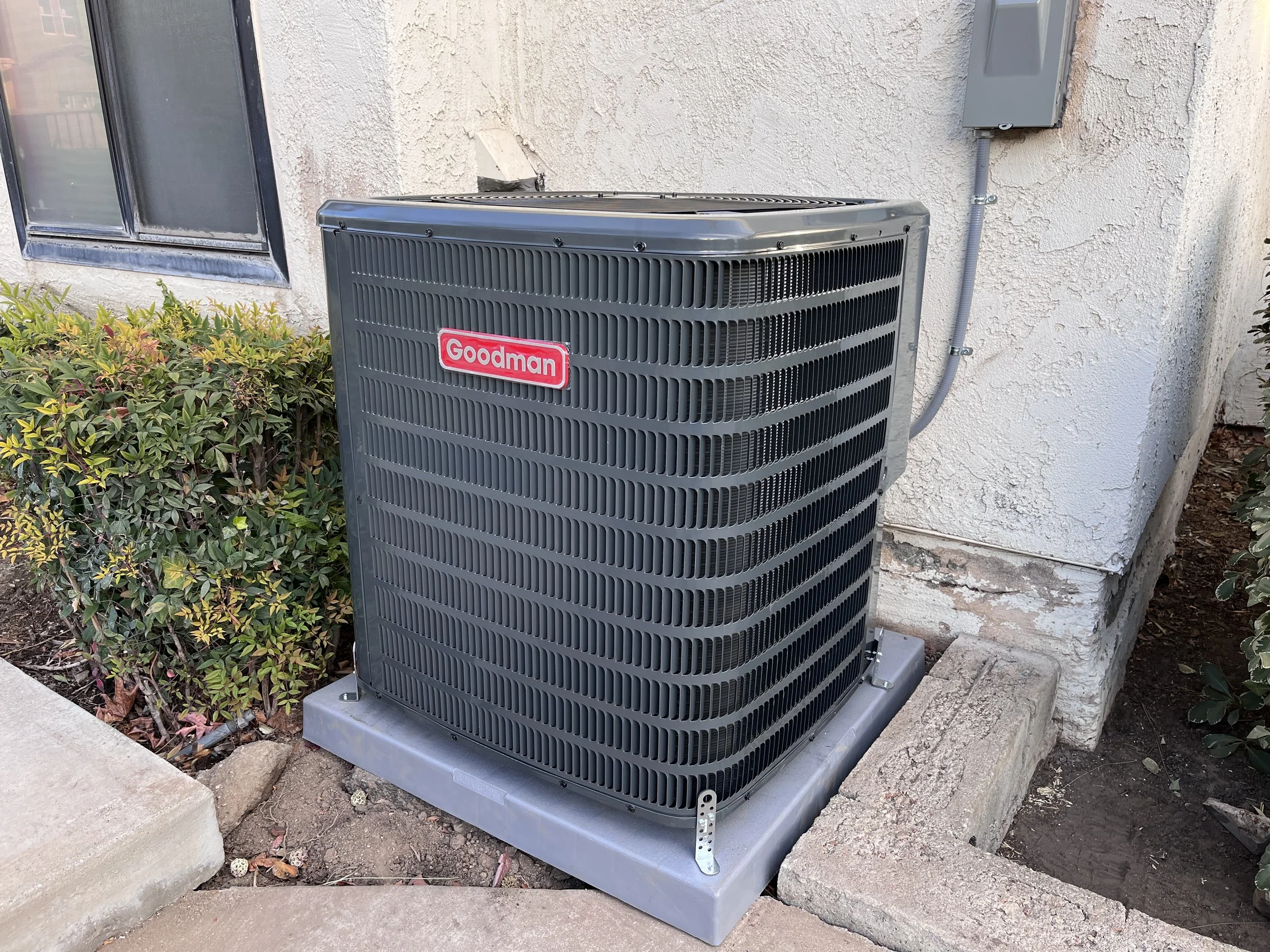 A Goodman brand air conditioning outdoor unit installed beside a house wall with a window, surrounded by landscaping including bushes and soil.