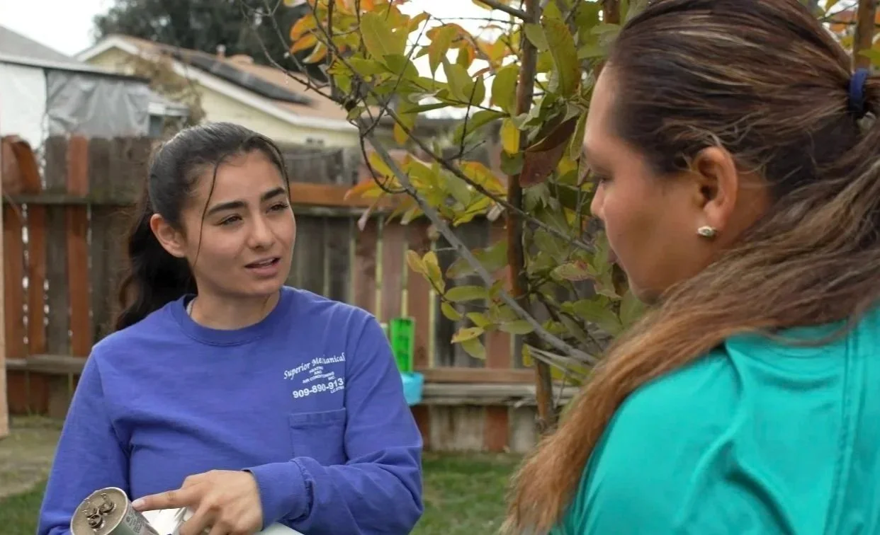 Two women having a conversation outdoors in a backyard. One woman, in a blue shirt, is speaking and holding a can; the other woman, in a teal shirt, is listening.