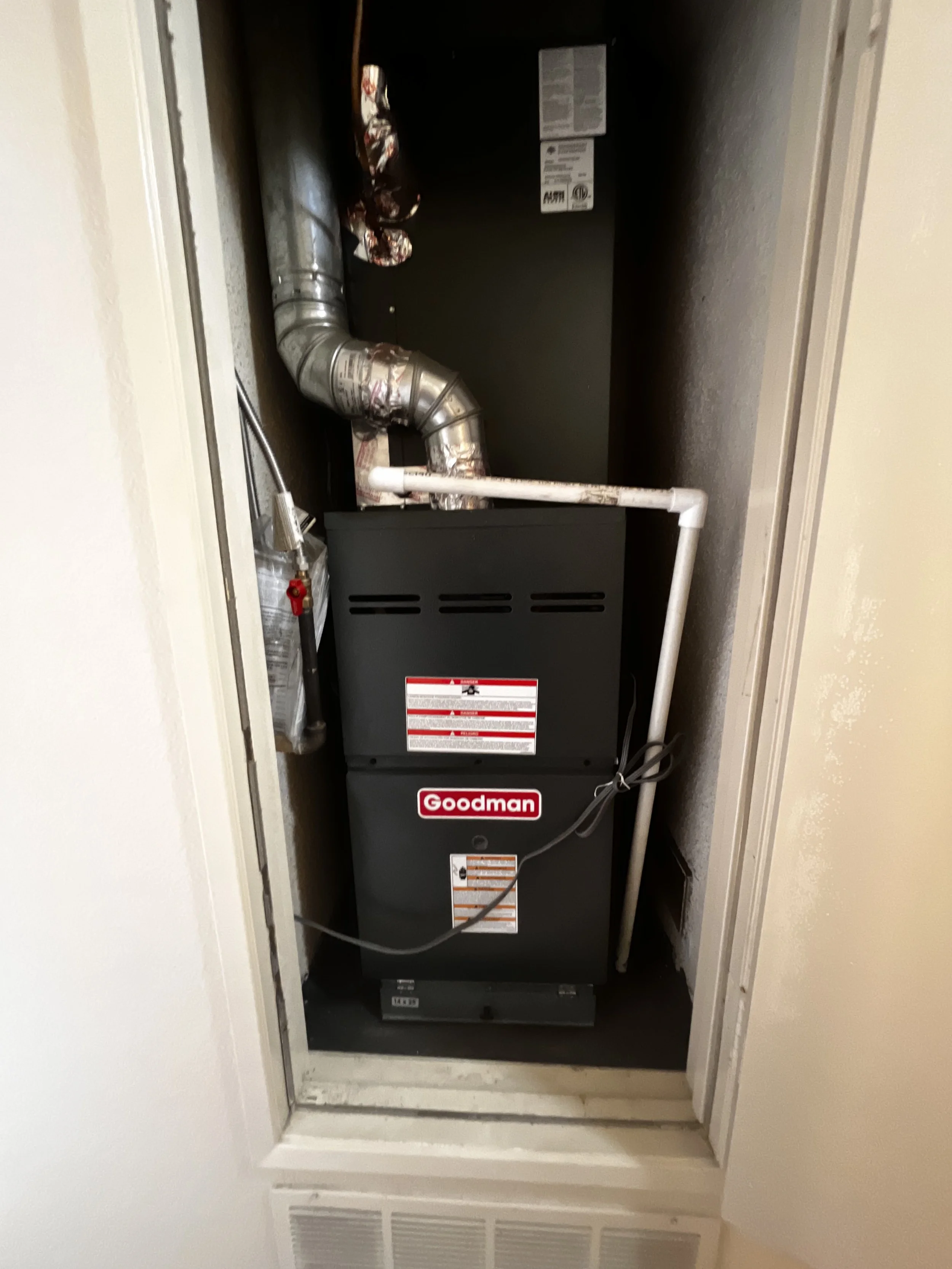 A Goodman furnace with ductwork and pipes installed in a small closet.