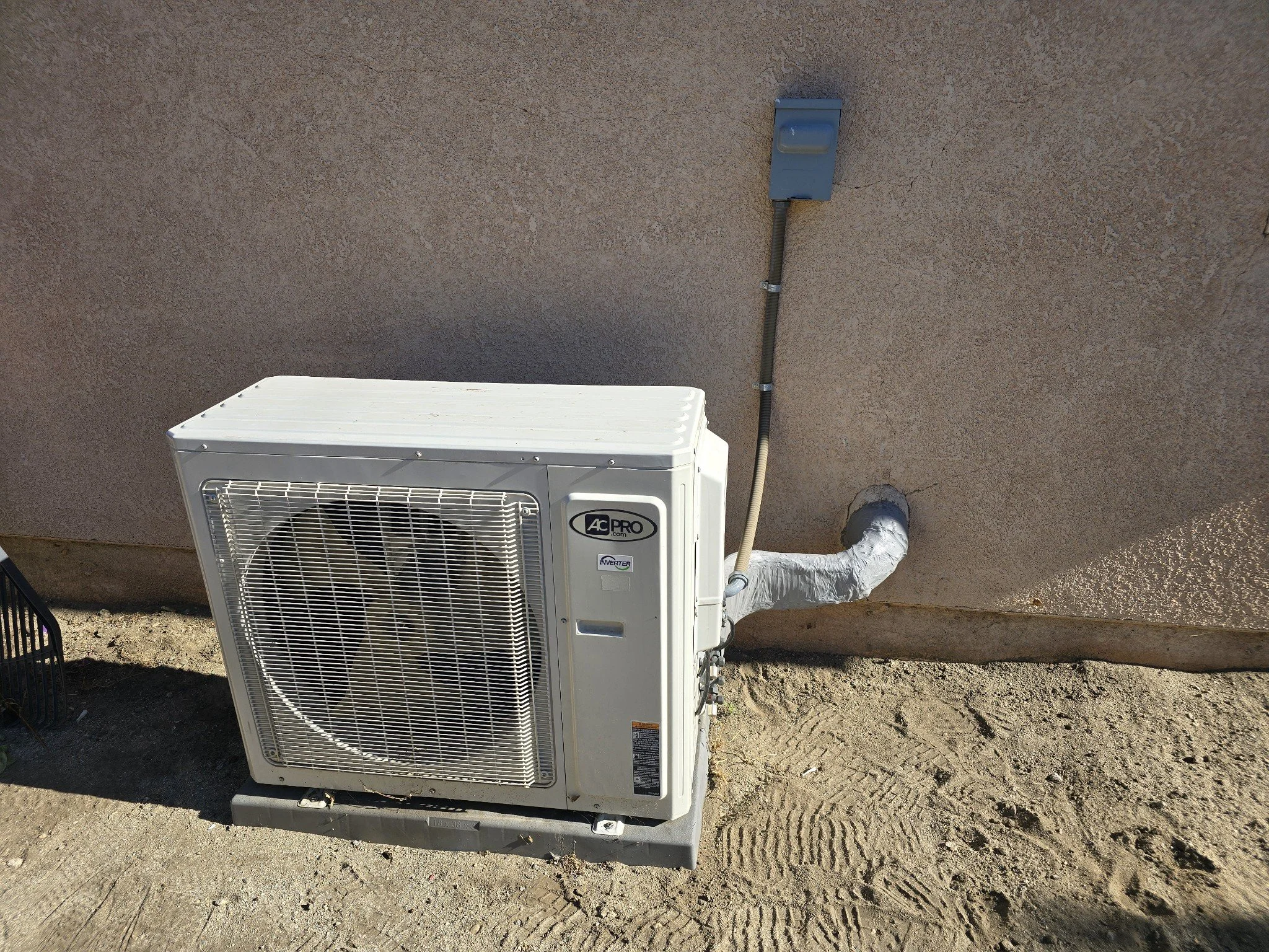 Outdoor air conditioning unit with a wall-mounted electrical box and conduit on a beige stucco wall, situated on a dirt ground.