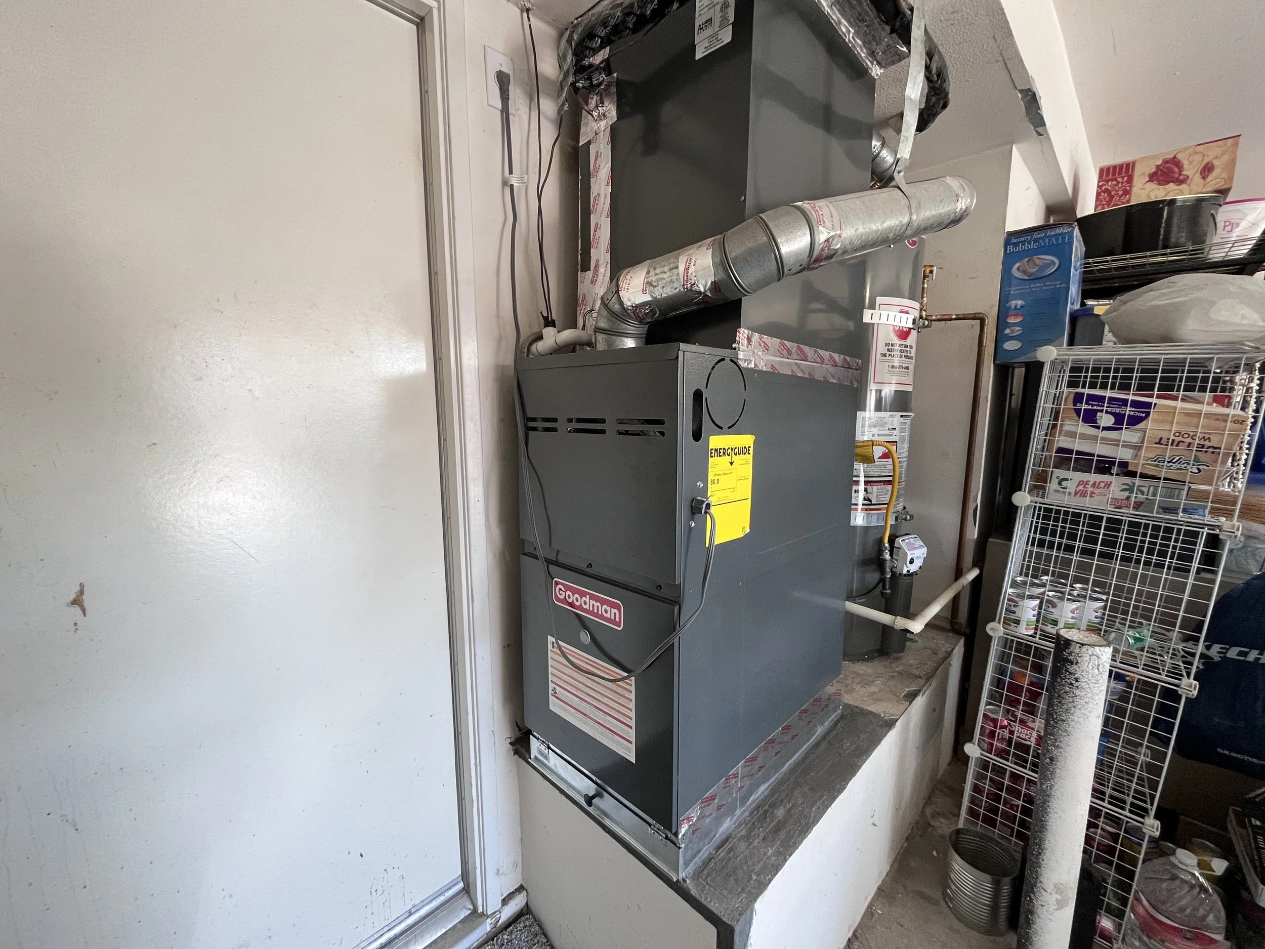 A Goodman HVAC furnace unit installed in a utility room next to a white wall and a sliding door, with a metal ventilation pipe and various electrical wiring visible.