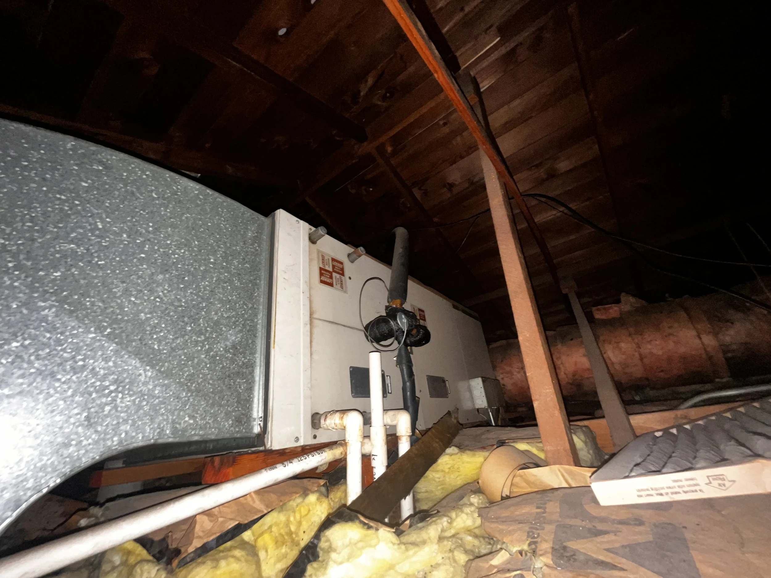 An HVAC system installed in an attic, with visible insulation and wooden rafters. Electrical wiring is also seen running along the ceiling.