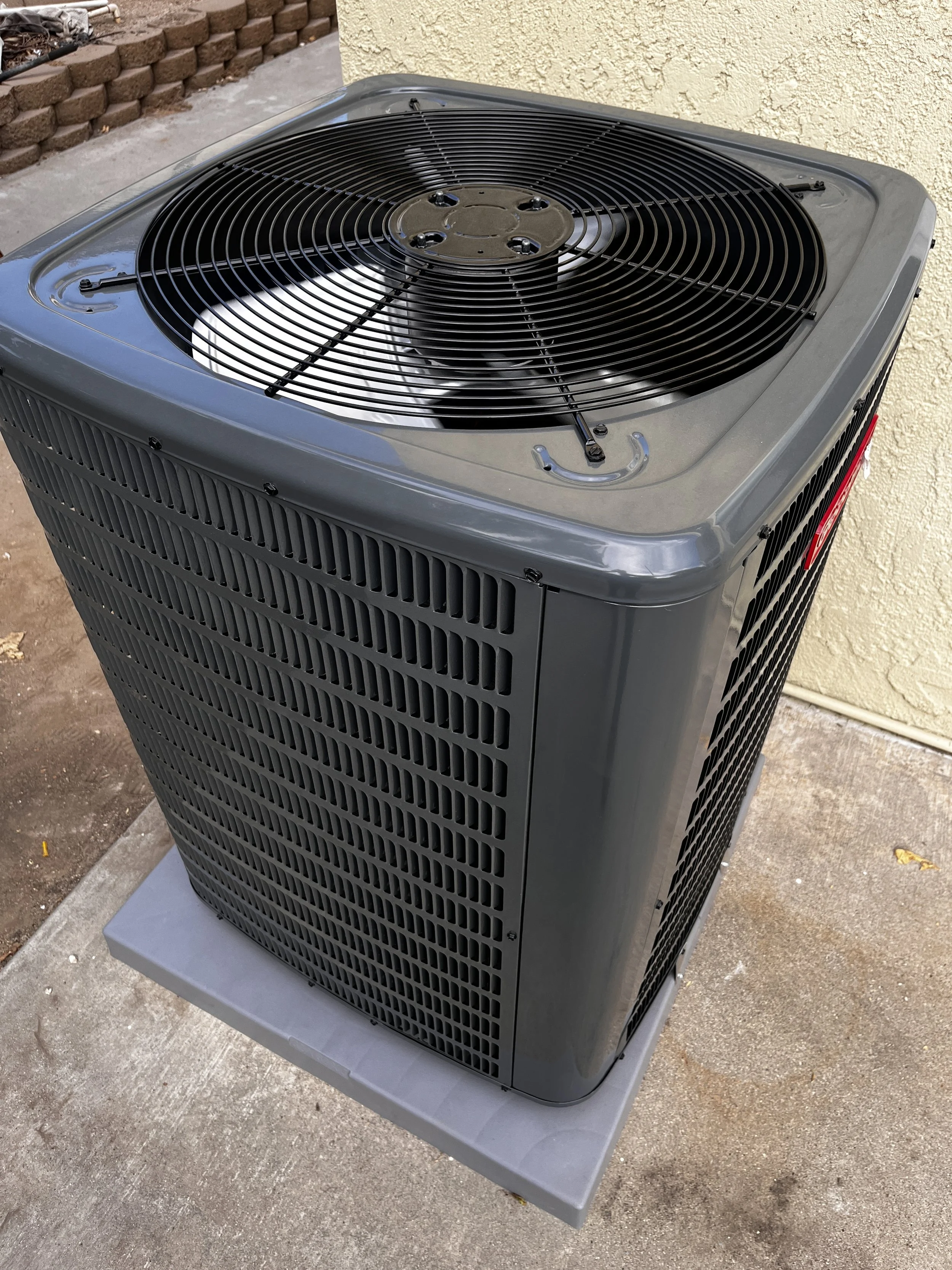 A gray outdoor central air conditioning unit with a large fan on top, situated on a concrete pad next to an exterior building wall.