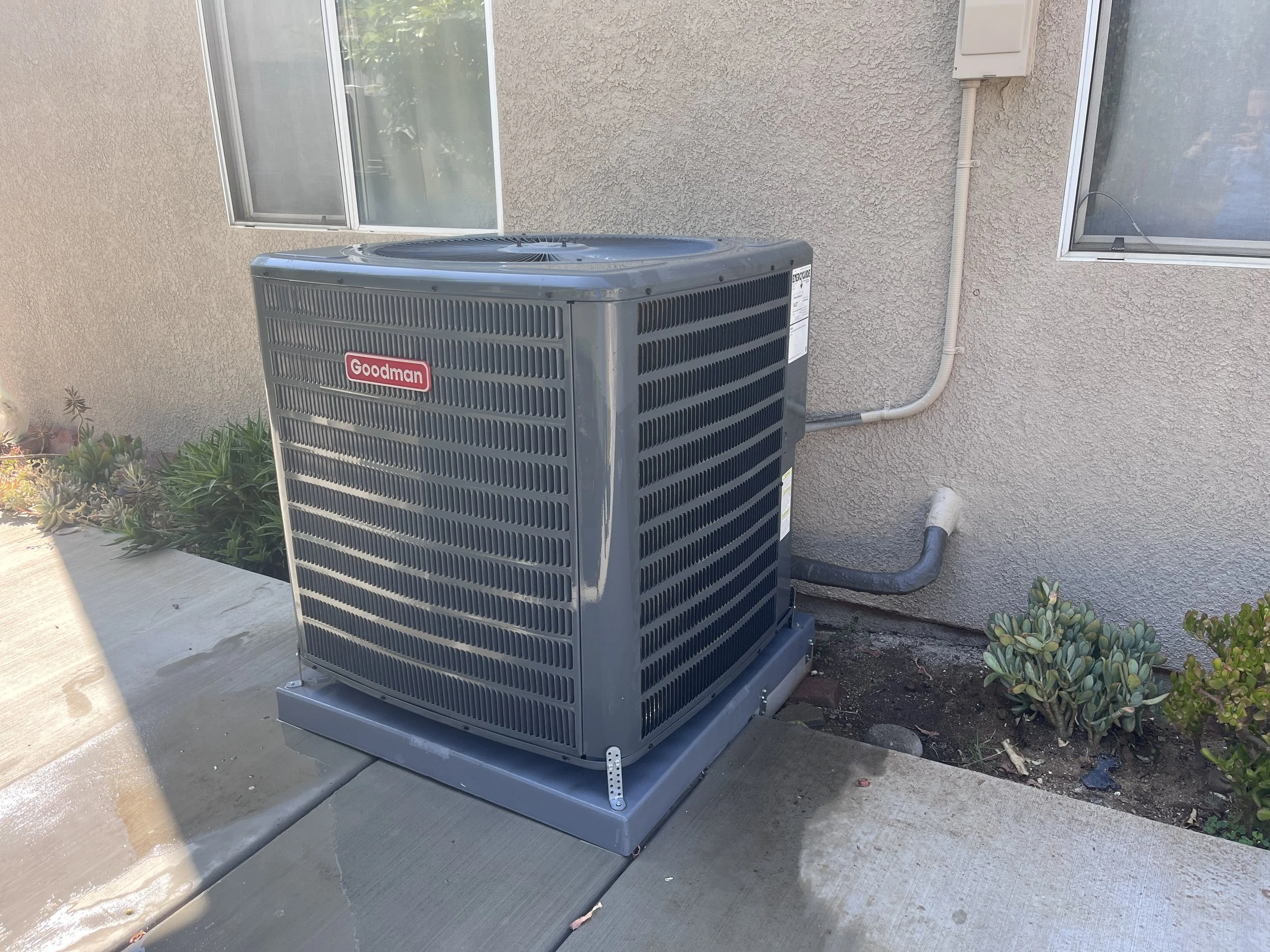 Outside air conditioning unit by the wall of a house with two windows and plants nearby.