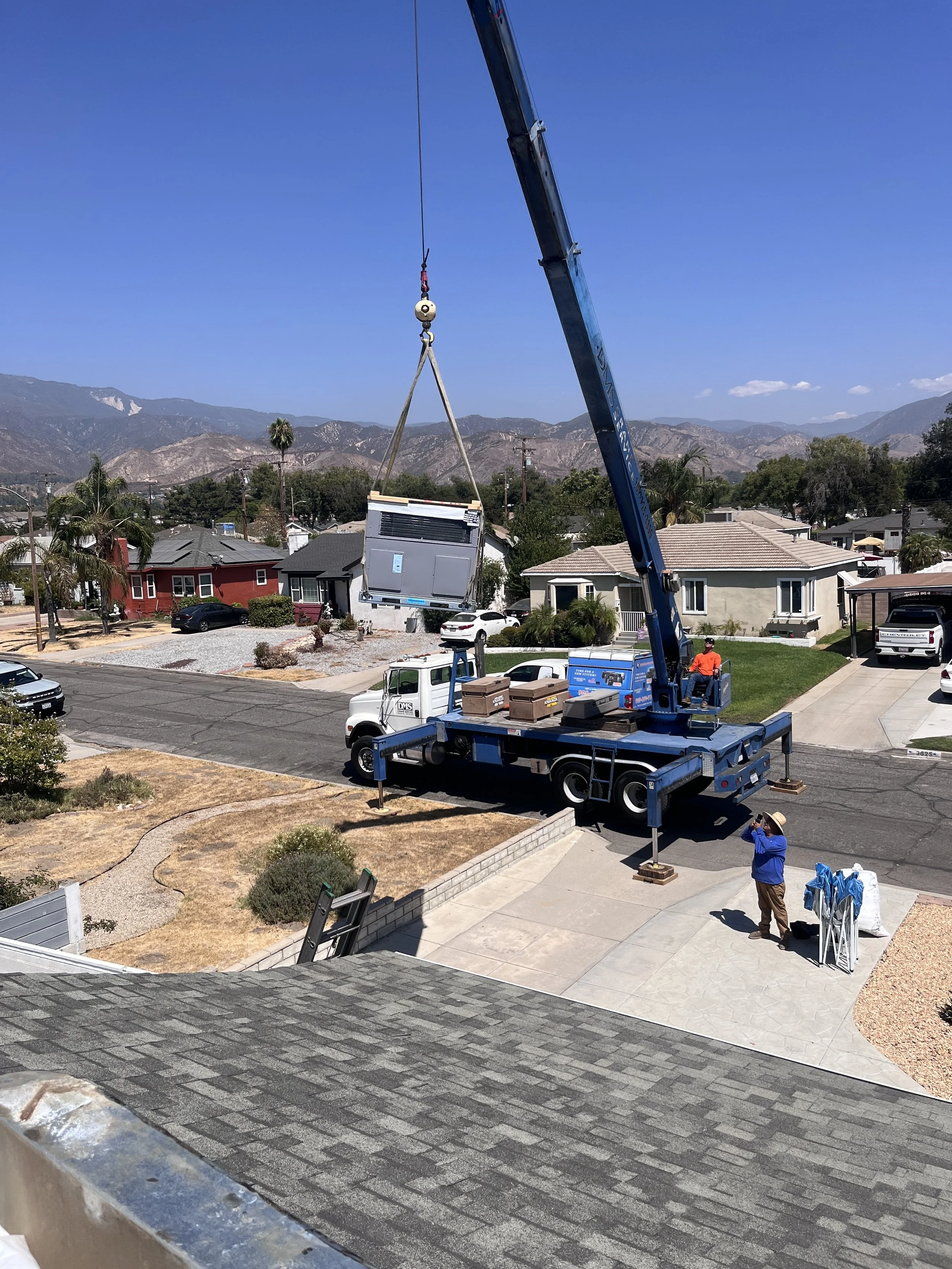A crane lifting a large air conditioning unit into a house yard, with a worker standing nearby on the sidewalk and another on the truck bed, set in a suburban neighborhood with mountains in the background.