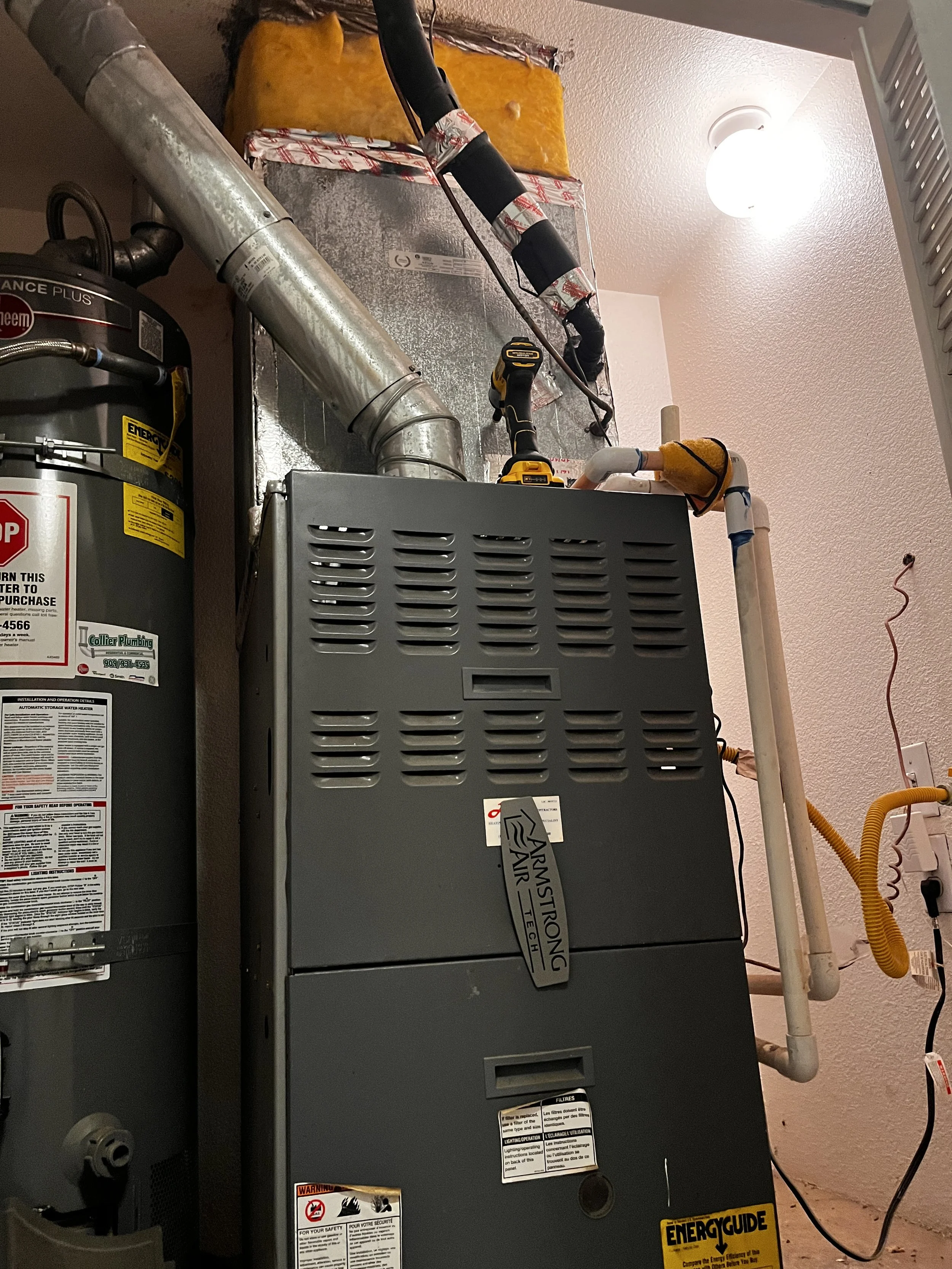 A close-up view of a furnace and water heater in a utility closet, with various pipes and tools around.