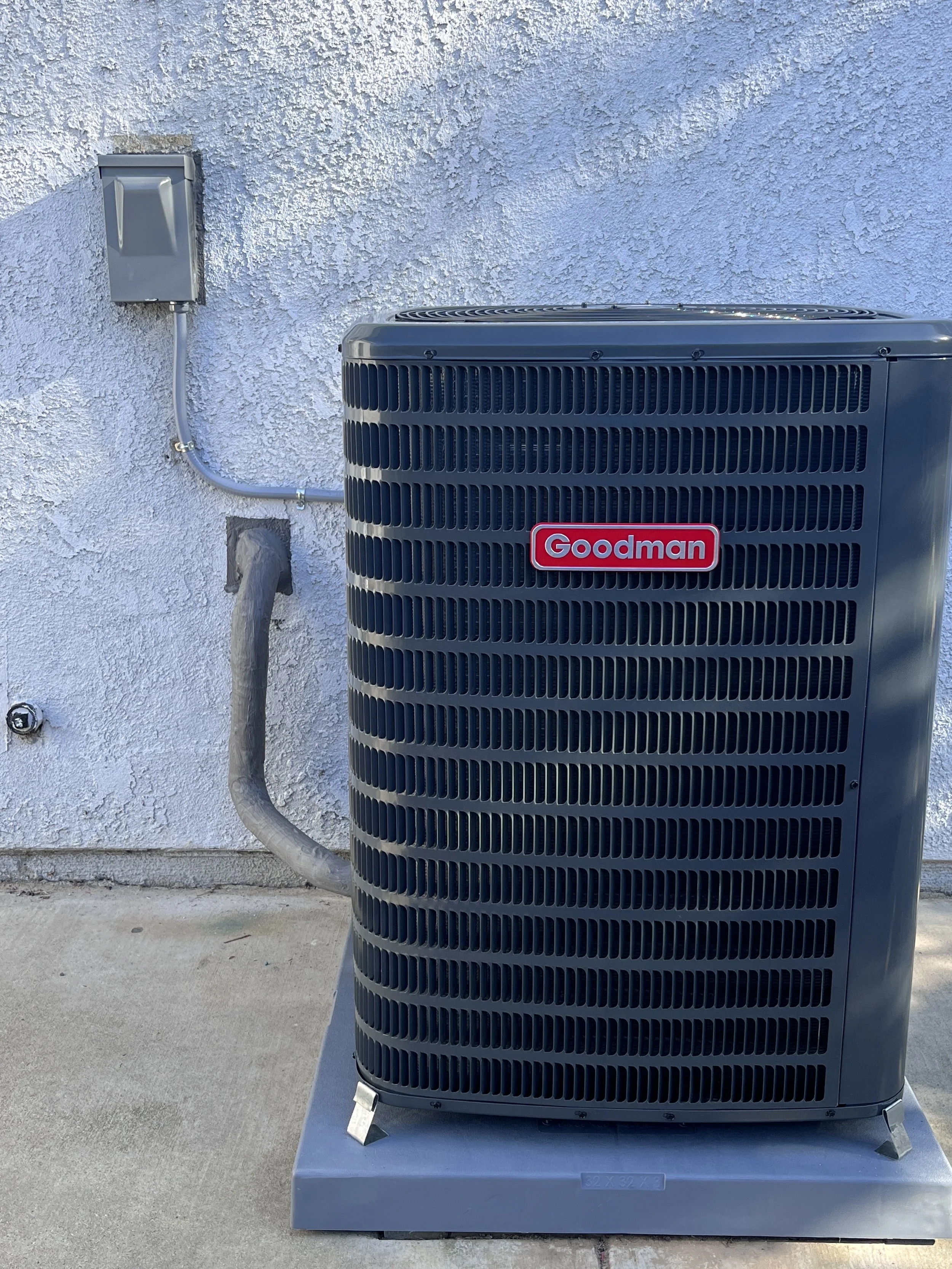 Exterior view of a Goodman air conditioning unit installed on a concrete pad against a textured white wall, with electrical connections and a vent pipe nearby.