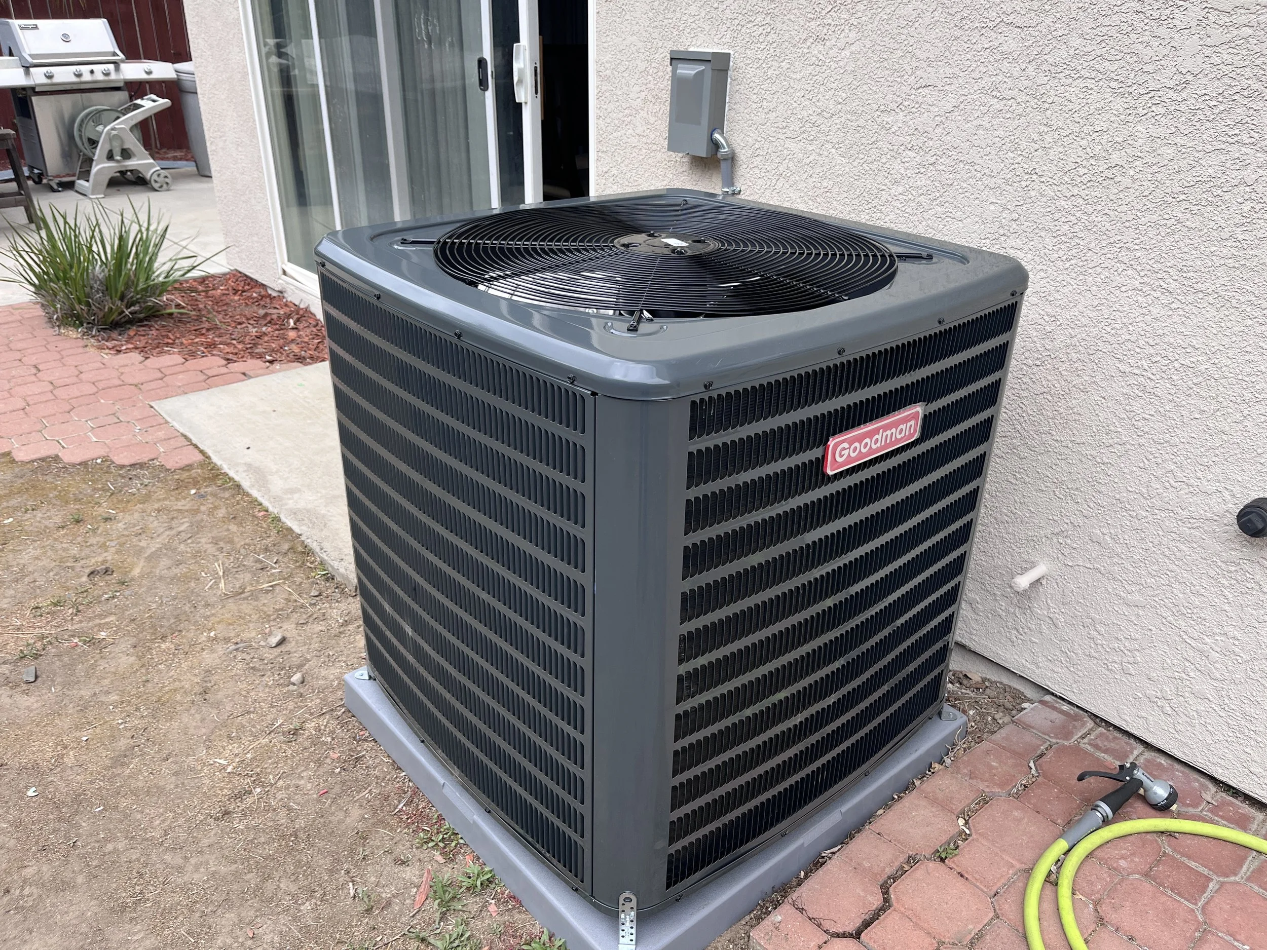 An outdoor air conditioning unit with a grille on top, placed on a raised platform near a beige stucco wall, next to a sliding glass door, with a yellow garden hose coiled nearby and red brick and concrete paving.