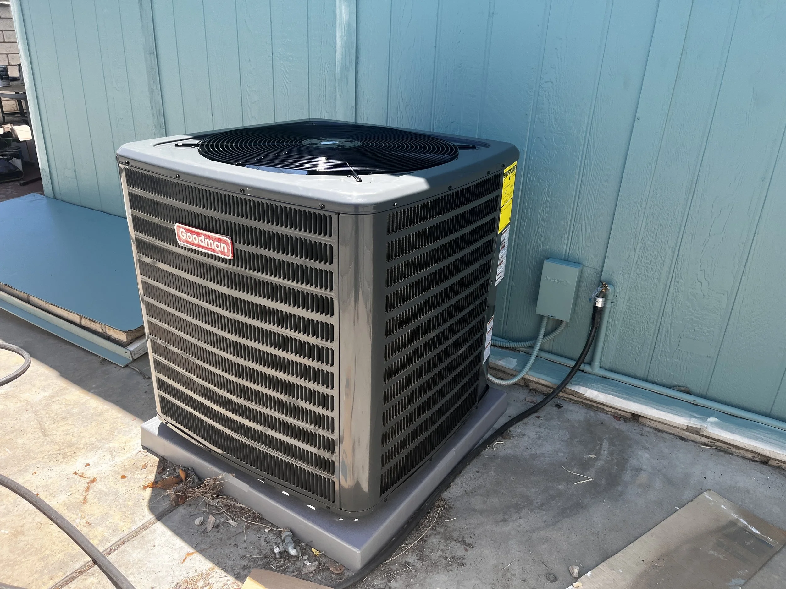 An outdoor Goodman air conditioning unit on a concrete platform next to a teal exterior wall of a building.
