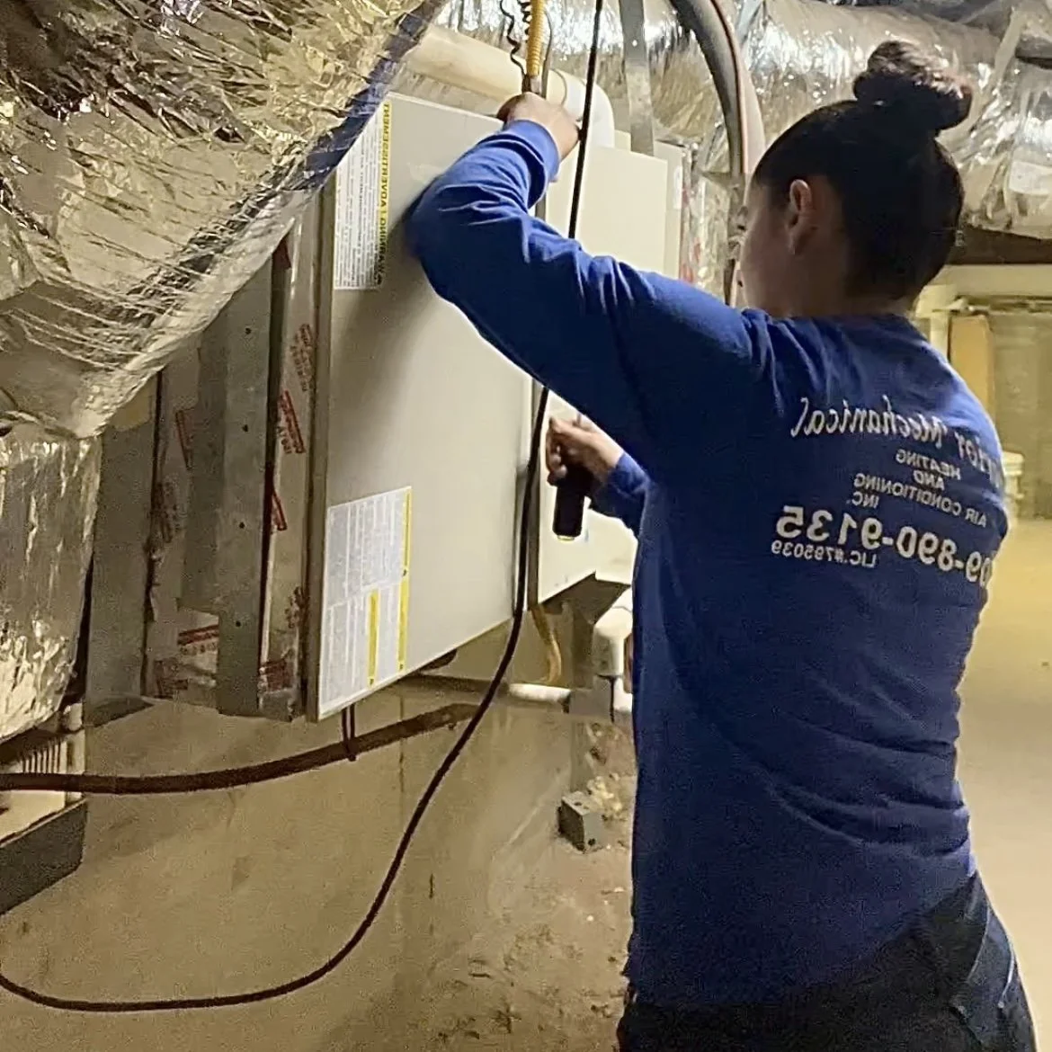 A technician repairing an HVAC unit in a basement, wearing a blue uniform and using a screwdriver.