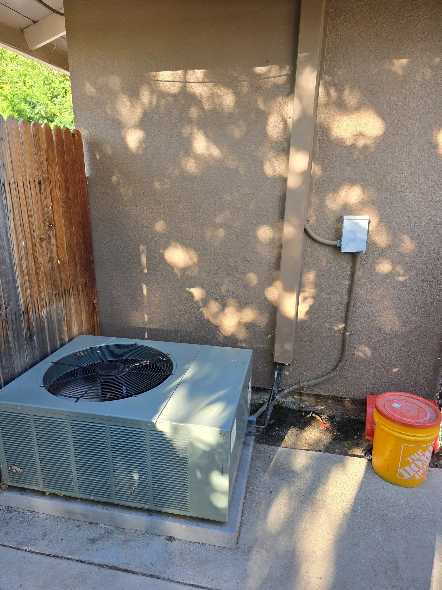 Outdoor air conditioning unit next to a beige wall, with a wooden fence on the left and a yellow Home Depot bucket on the right. Shadows of leaves are cast on the wall.