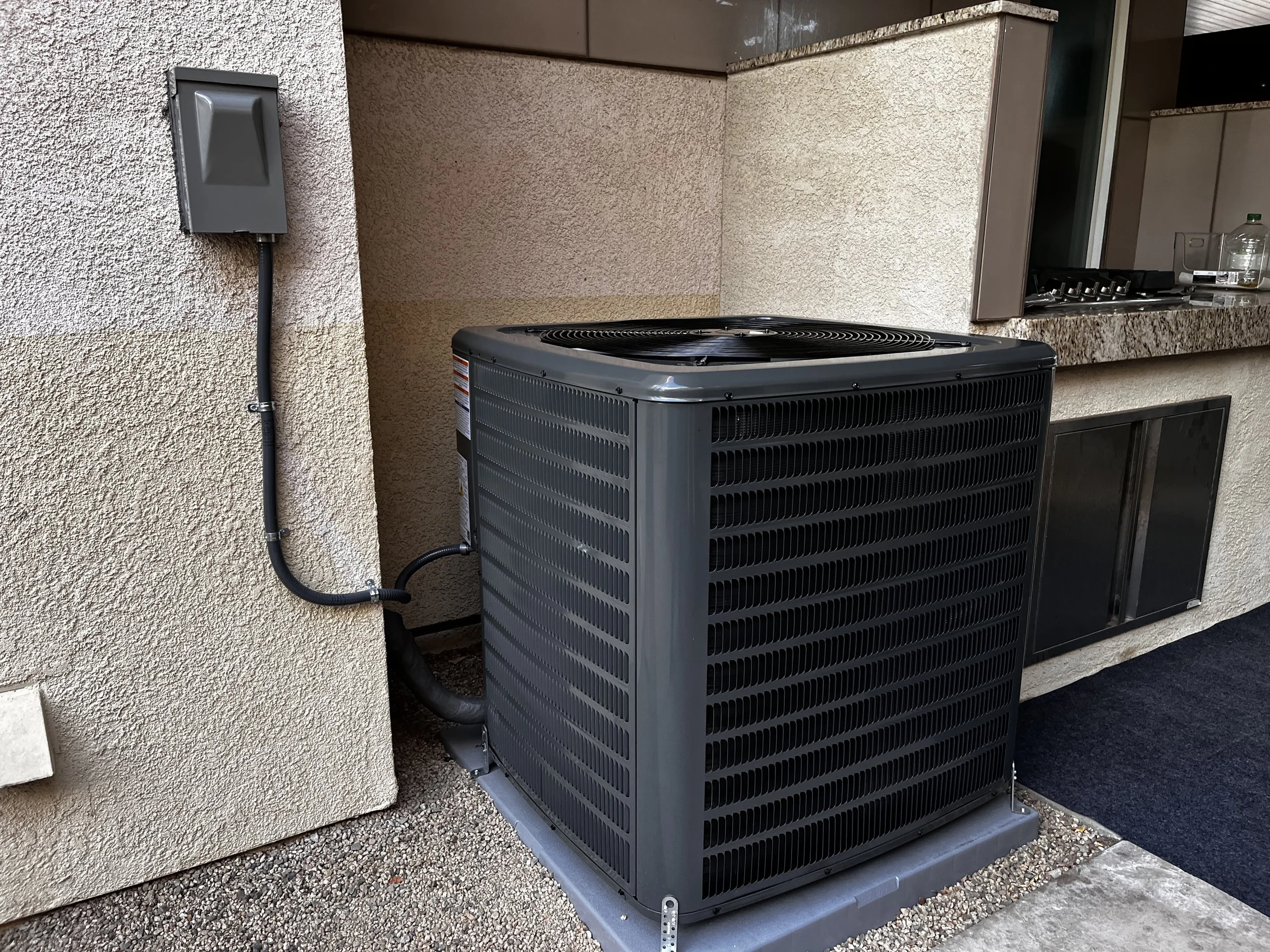 Outdoor air conditioning unit on a concrete pad against a house wall with a black drain pipe and an electrical box.