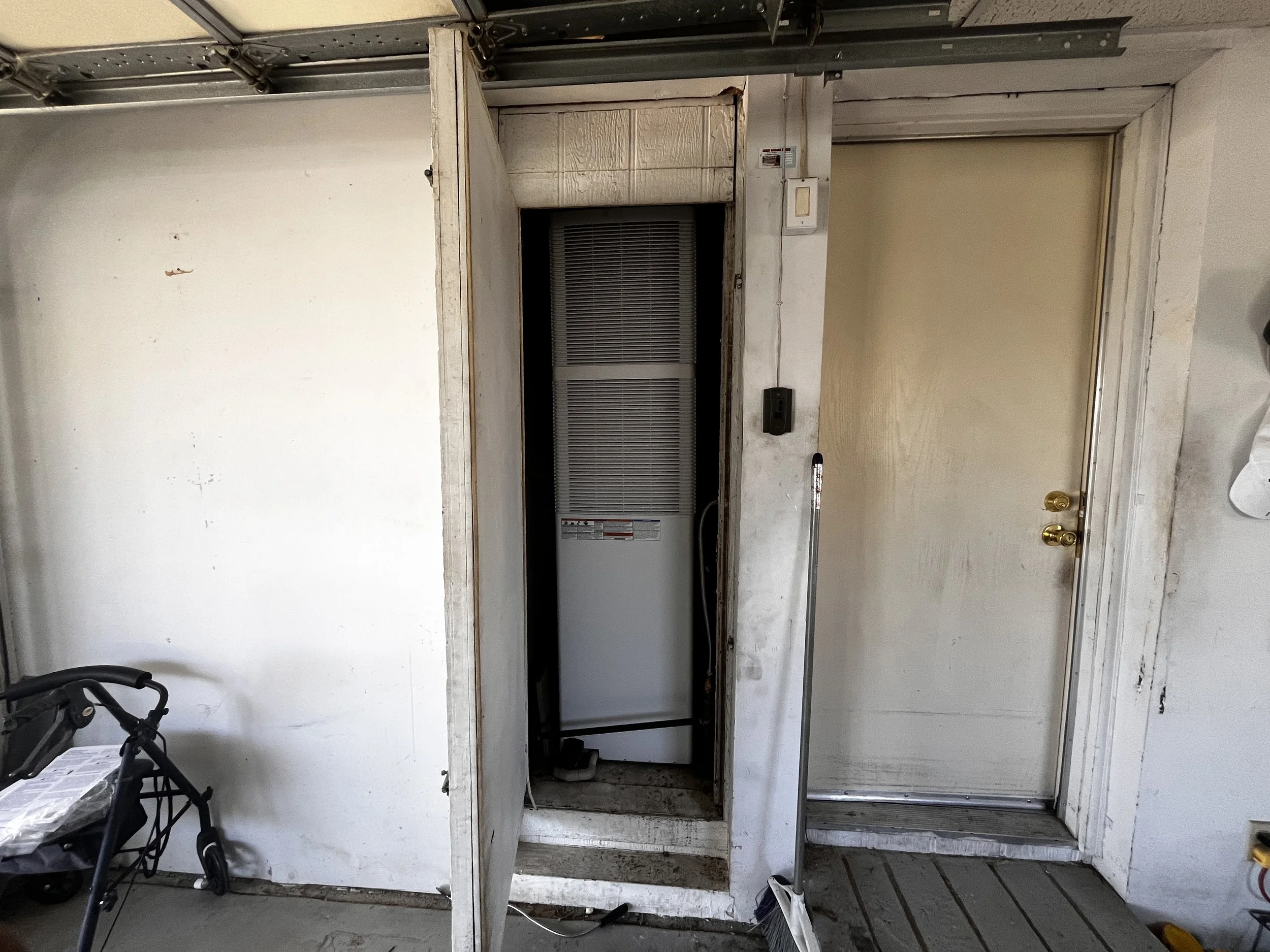 A utility room with a closed white door on the right, and a small open closet or electrical panel on the left, with an HVAC unit inside. The area appears to be in a garage or basement with a concrete floor and exposed walls.