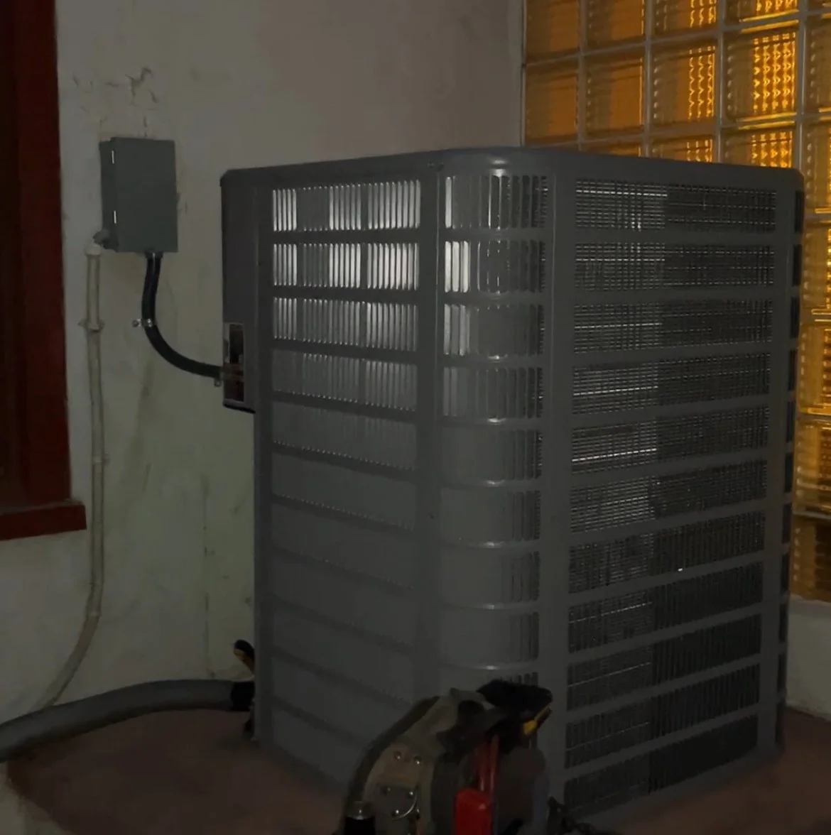 An industrial air conditioning or cooling unit installed on a wall in a room with a yellow-tinted glass block window.