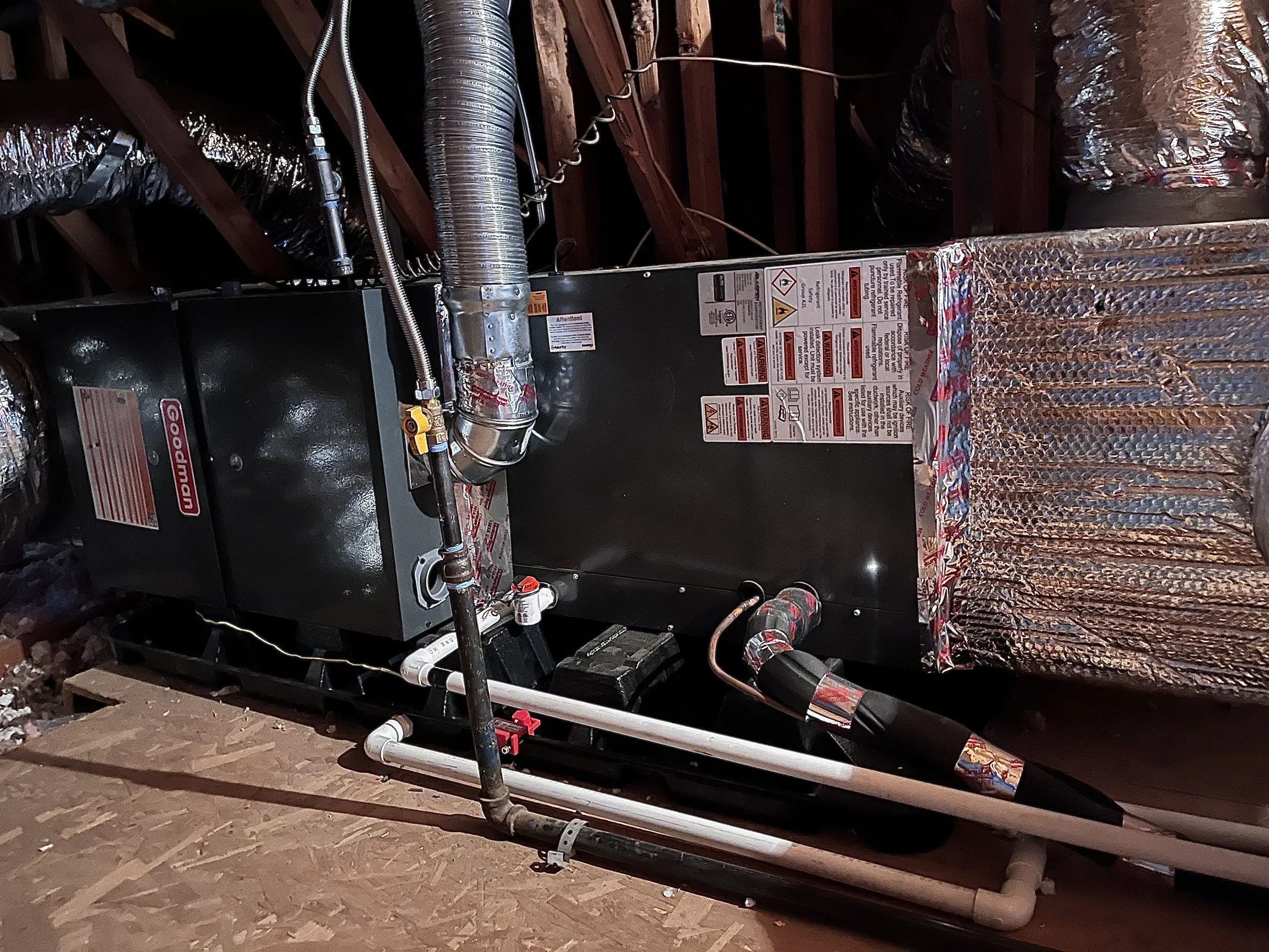 An HVAC furnace unit installed in an attic, with visible ductwork, pipes, and informational labels on the black metal surface.