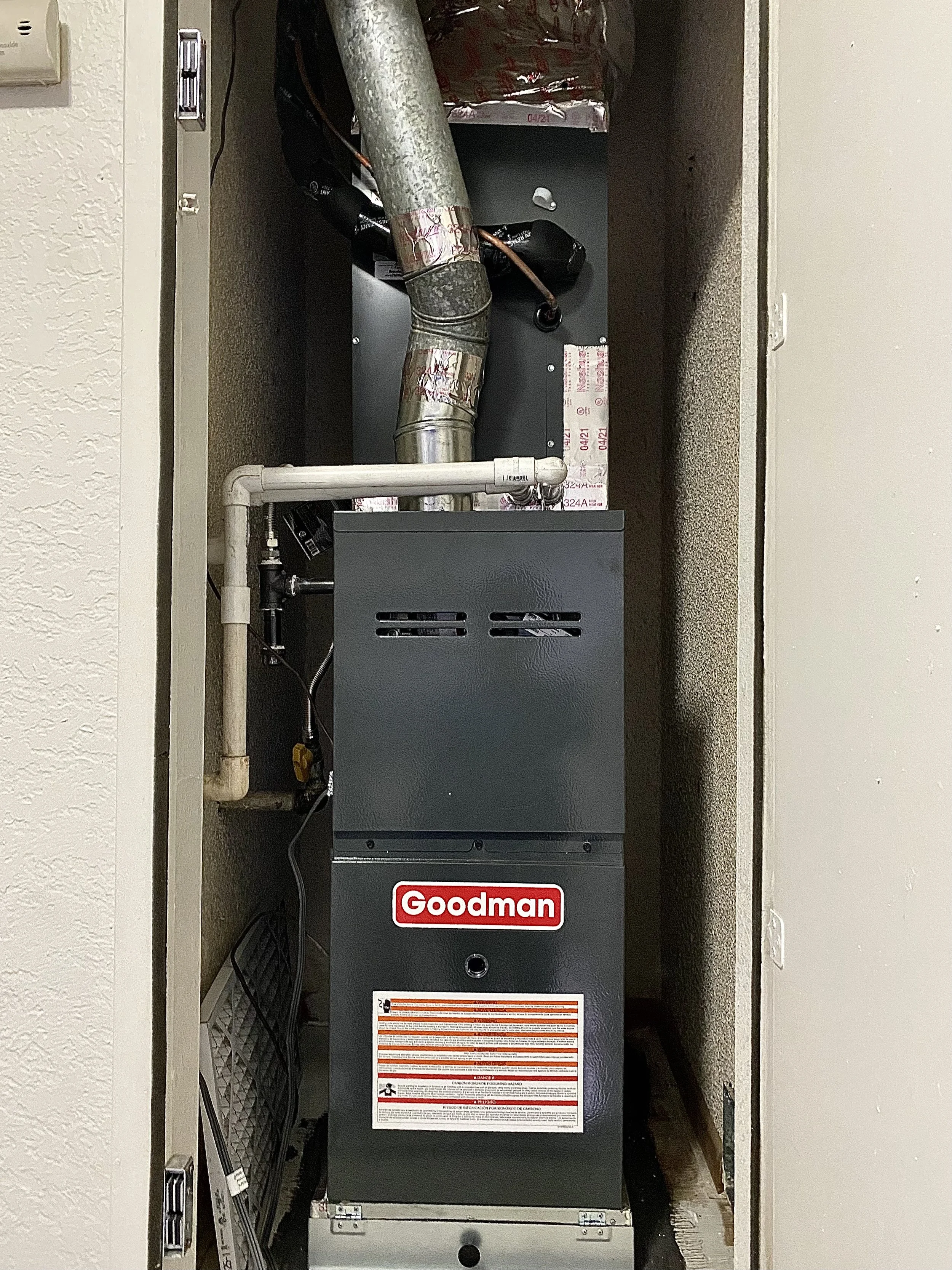 A Goodman HVAC furnace system inside a small closet with pipes and ducts.