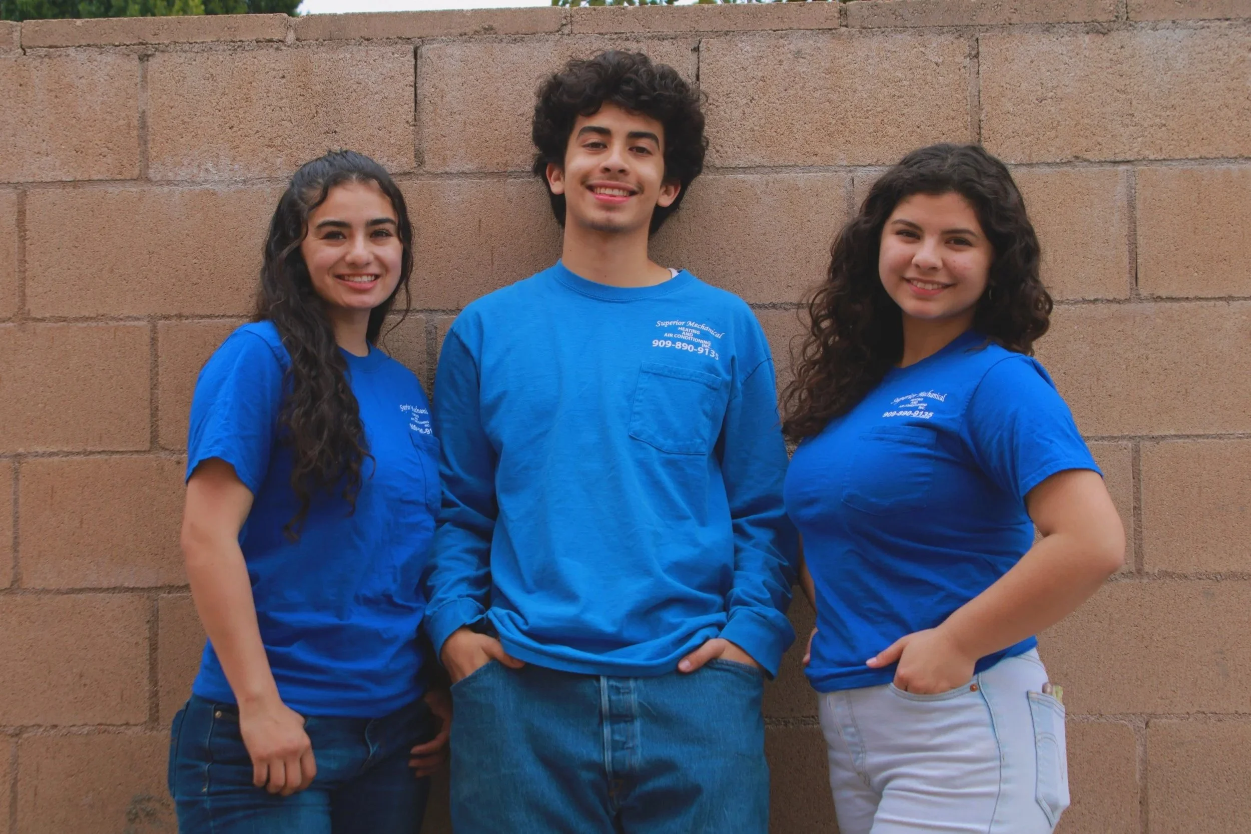 Three young people wearing blue work shirts stand against a beige brick wall, smiling at the camera.