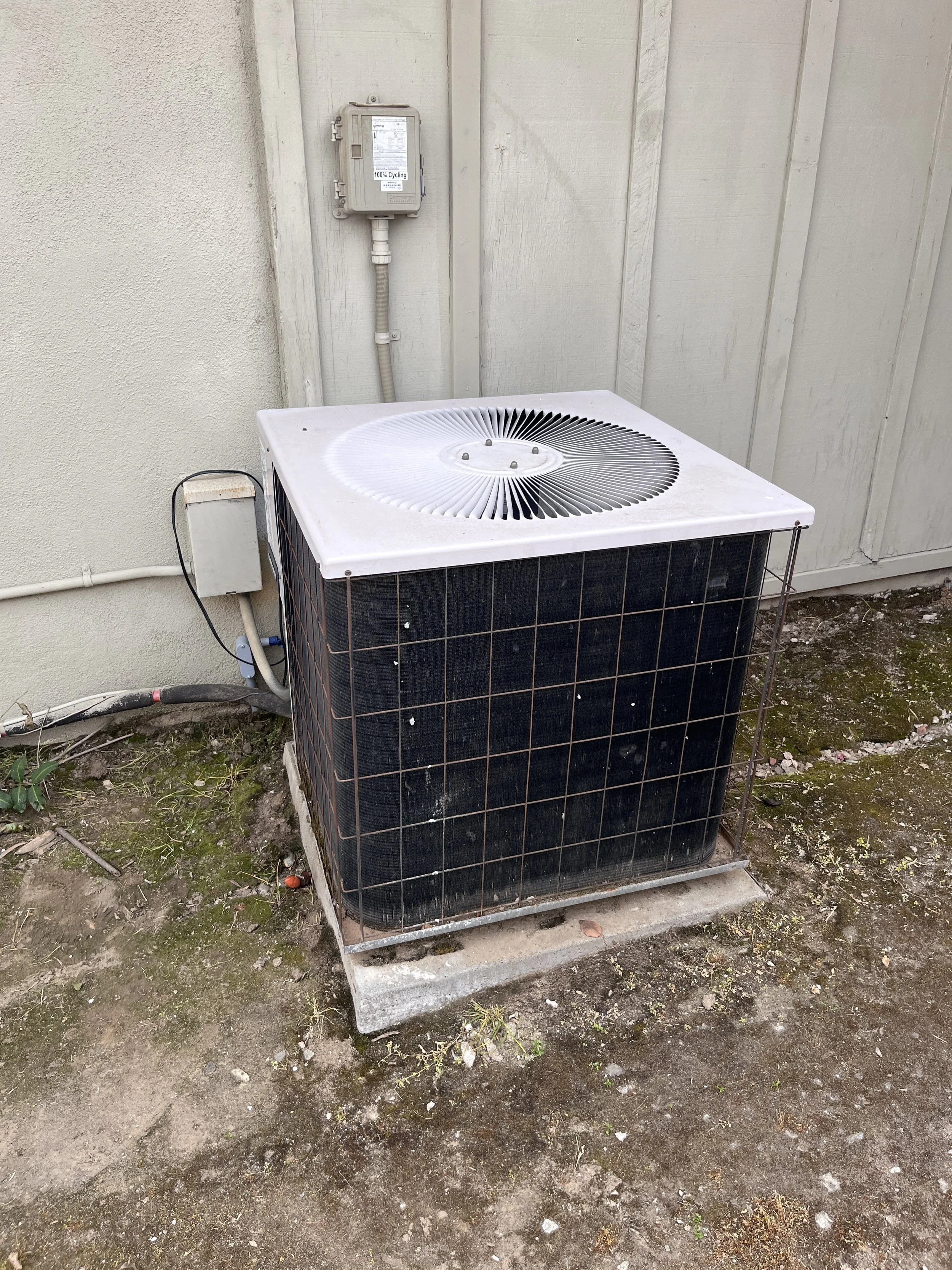 An outdoor air conditioning unit on a concrete slab next to a beige wall, with a white electrical box and electrical meter mounted on the wall.