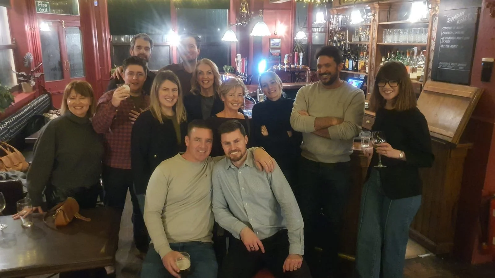A group of people gathered in a bar or pub, smiling and posing for a photo, some holding drinks.