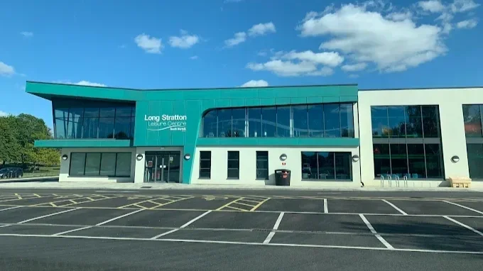 Exterior of the Long Stratton Leisure Centre, a modern building with a teal and white facade, large glass windows, and an empty parking lot in front.