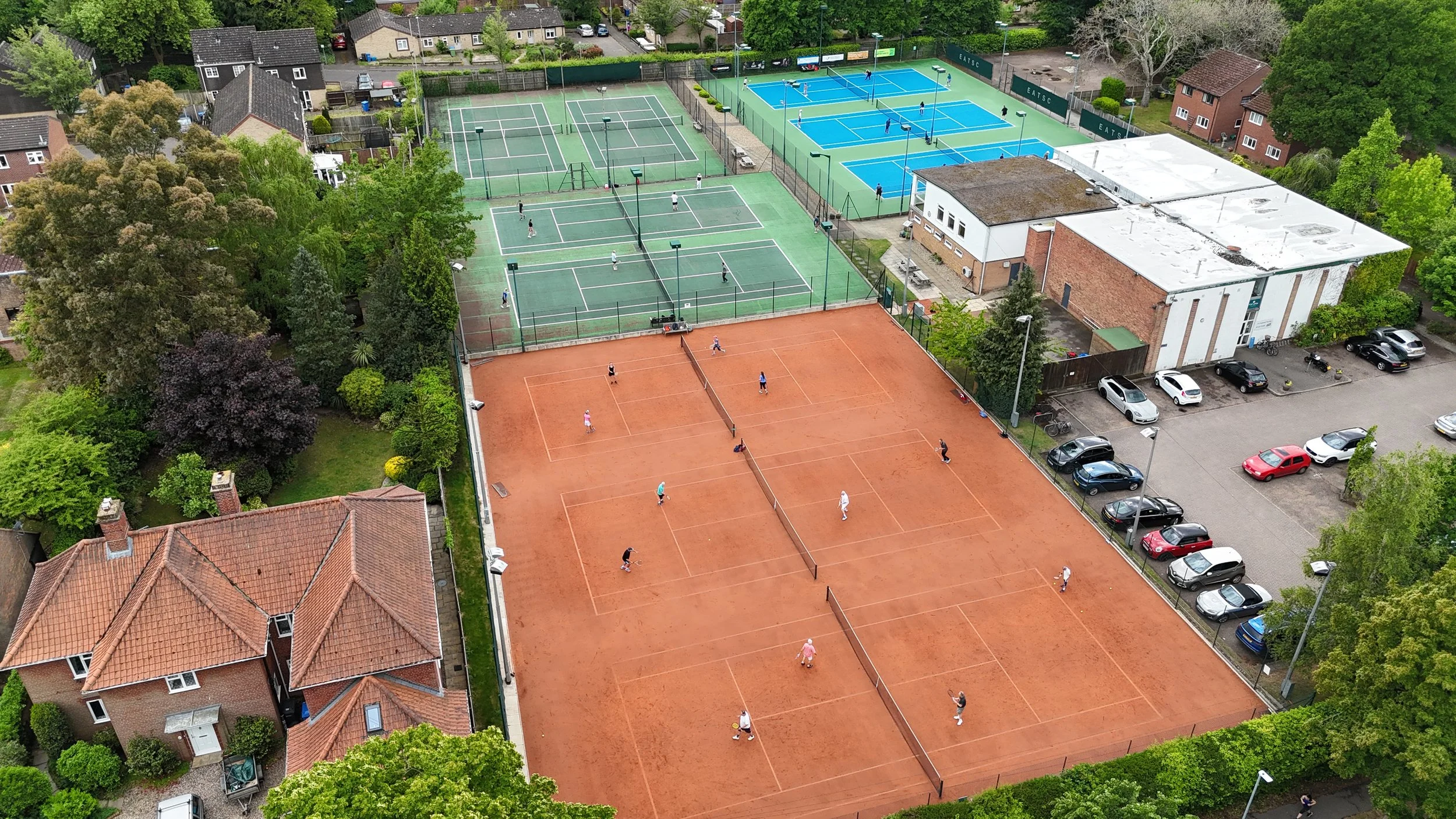 Aerial view of a sports complex with multiple tennis courts, some are clay and others are hard courts, with people playing tennis, surrounded by residential houses and parking lot with cars, trees, and green foliage.