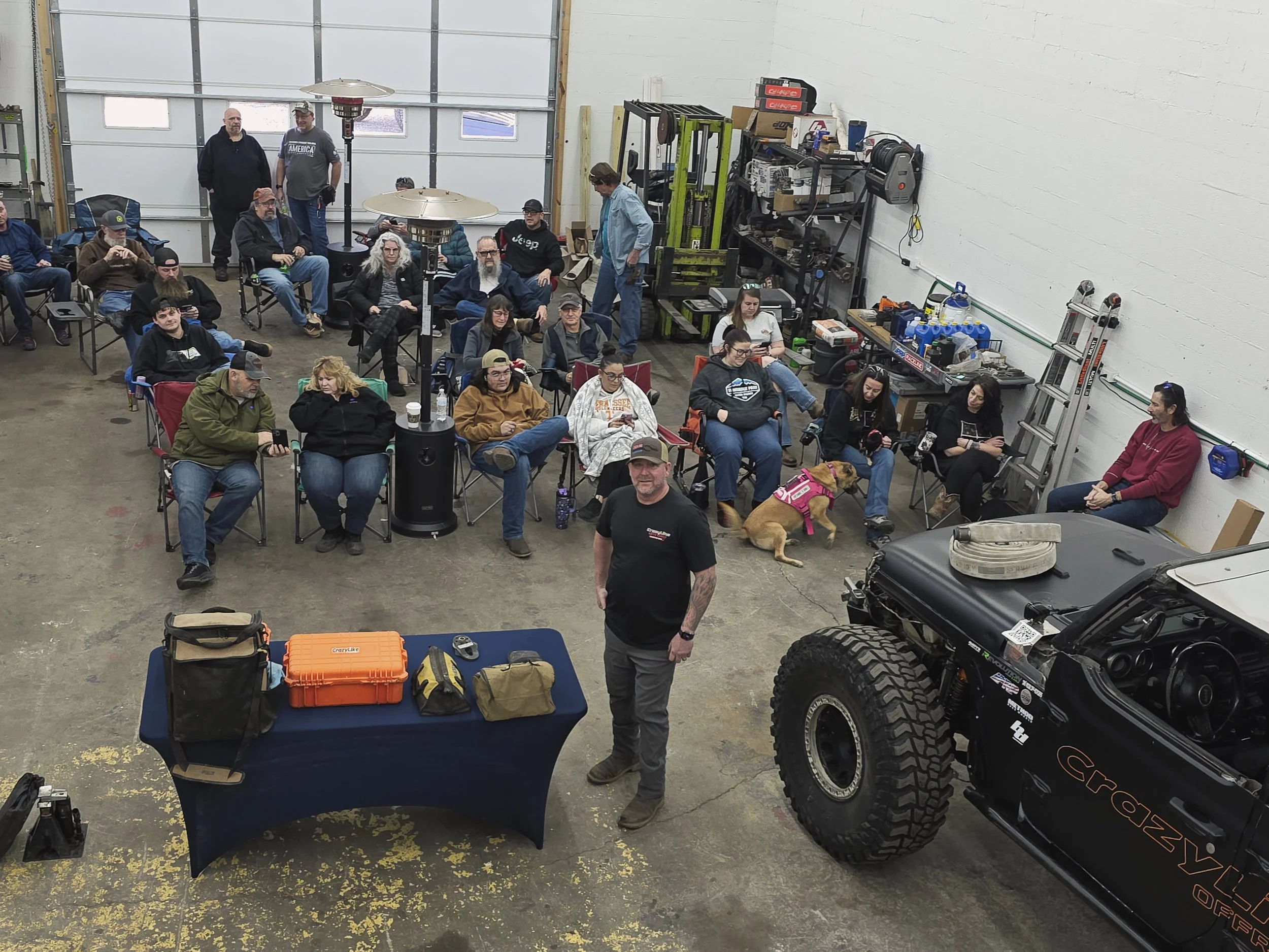 People seated in a workshop or garage with a man standing in front of a table with equipment, and a large off-road vehicle on the right.