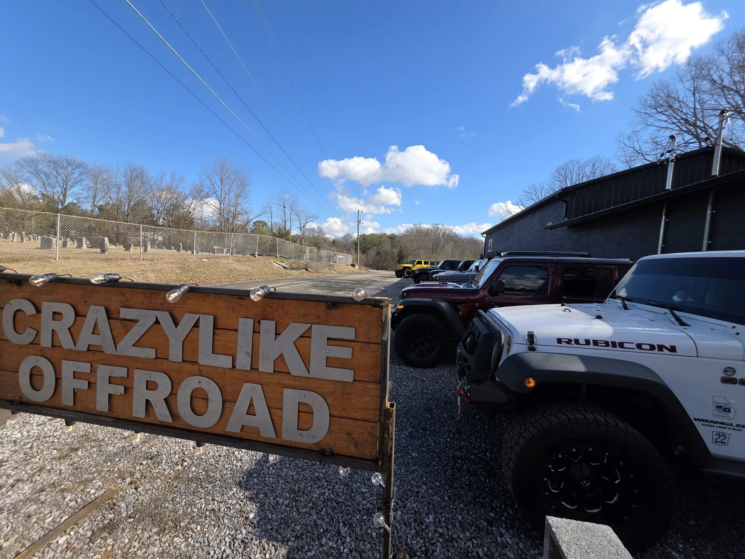 A wooden sign with the text "CRAZYLIKE OFFROAD" hangs on a chain-link fence, surrounded by parked off-road vehicles, including a white Jeep Rubicon and a maroon vehicle, with a building and a hill with trees in the background under a partly cloudy sky.